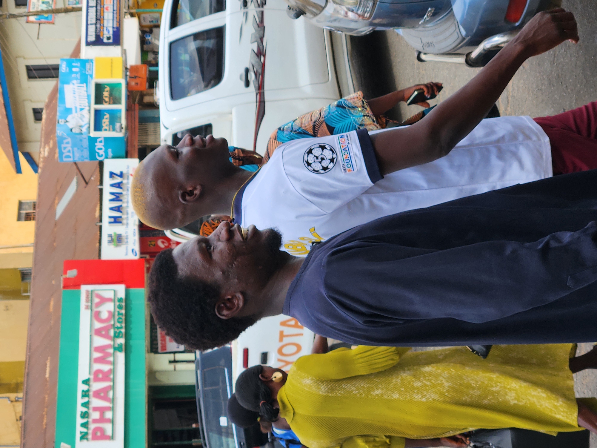 The image captures a street scene in the city of Unknown, Unknown. Two men are visible in the foreground: the first is wearing a white shirt with a soccer-related design and a gold necklace, while the second is wearing a dark blue shirt. A woman wearing a yellow dress is also visible. Behind them, a white Toyota vehicle is parked. Various businesses line the street, including "Nasara Pharmacy & Stores" and "HAMAZ." There are also signs for "DStv" and "GOtv". Pedestrians are walking along the street.