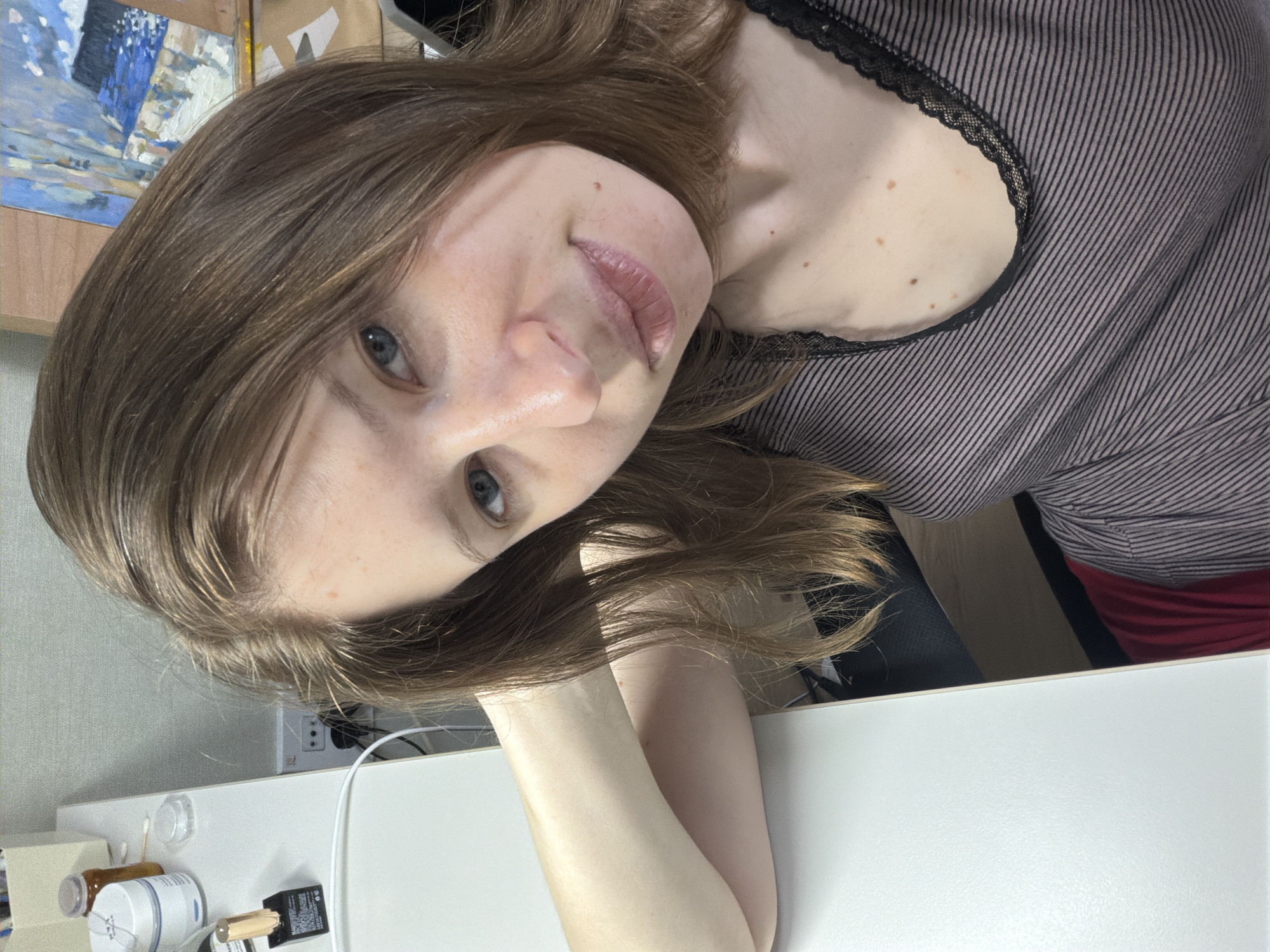 A young Caucasian woman with long, brown hair is shown in a close-up, overhead shot. She is sitting at a white desk, her left arm propped up and resting behind her head. Her eyes are open and she is looking towards the camera with a neutral expression. The woman is wearing a striped top with lace detailing at the neckline. On the desk to her left are various cosmetic items, including a jar of cream, a bottle of nail polish, and a cotton swab. In the background, an easel with a painting on it is visible. The scene appears to be an indoor setting, likely a home or studio, with no specific identifying landmarks of Dongducheon-si, South Korea.