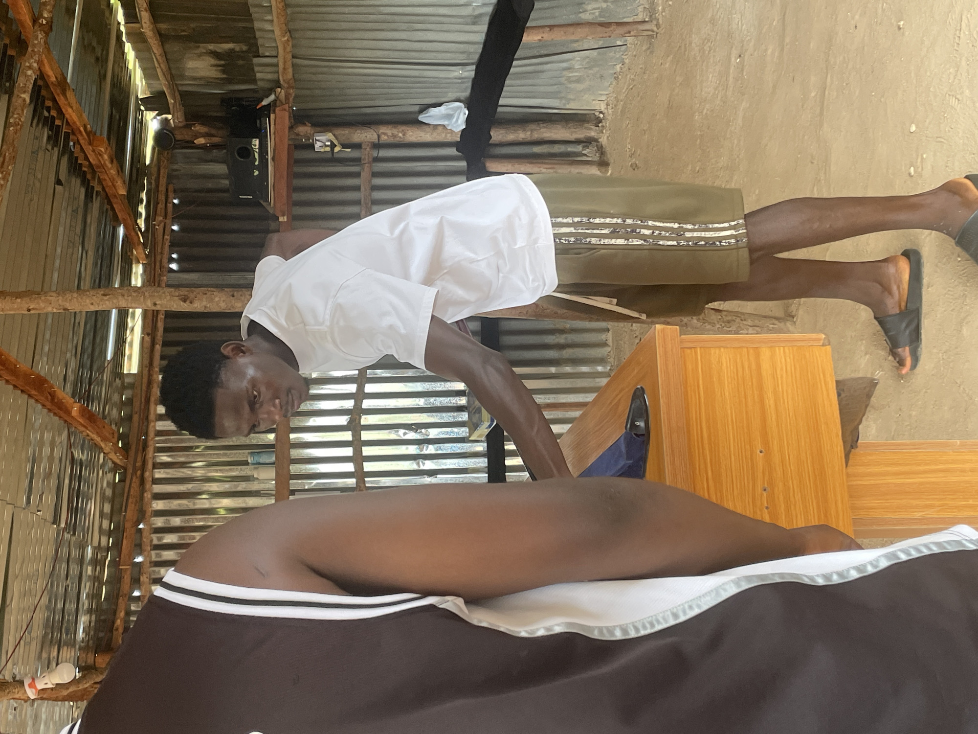 In Durimi, Nigeria, a young Black male, dressed in a white t-shirt and green shorts, is performing a massage on another individual lying on a bed covered with a brown and white sheet. The setting appears to be an outdoor or semi-outdoor structure with a corrugated metal roof supported by wooden beams. The background shows a rustic environment with the textured appearance of dirt or concrete on the ground. A wooden cabinet or table is visible to the right of the frame. The lighting suggests it is daytime. The individual receiving the massage has dark skin and is partially visible, lying on their back. The masseur's face shows concentration as he works.