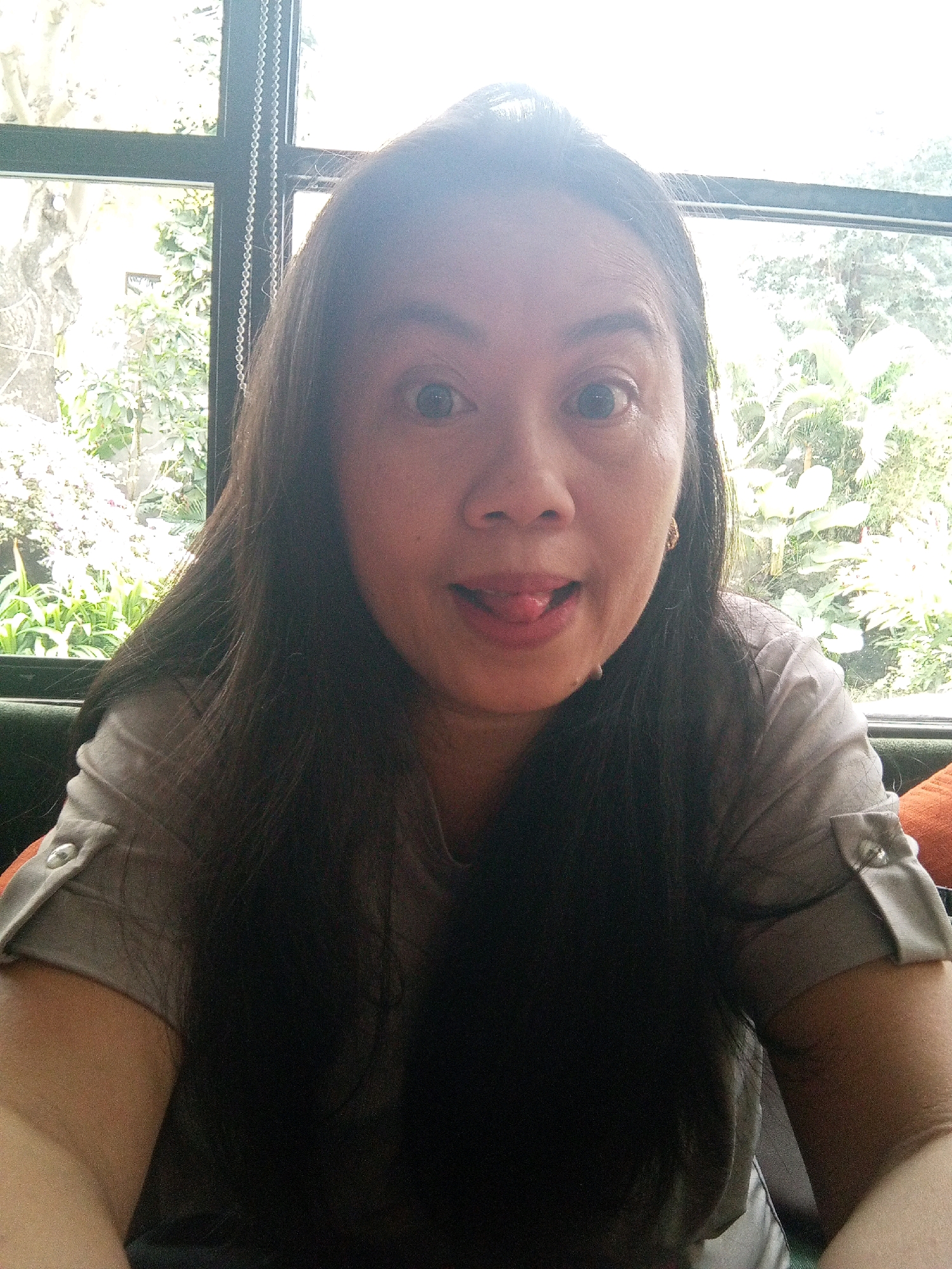 A woman is taking a selfie in front of a window. She has long, dark hair and is sticking her tongue out. She is wearing a grey t-shirt with rolled-up sleeves. Behind her, a window shows a lush green garden with trees and plants. The woman is likely in a restaurant or cafe, given the seating and the window view. The sunlight is coming from the window, creating a bright, natural light. The location of Denpasar, Indonesia is not discernible from the image itself; it is inferred information.