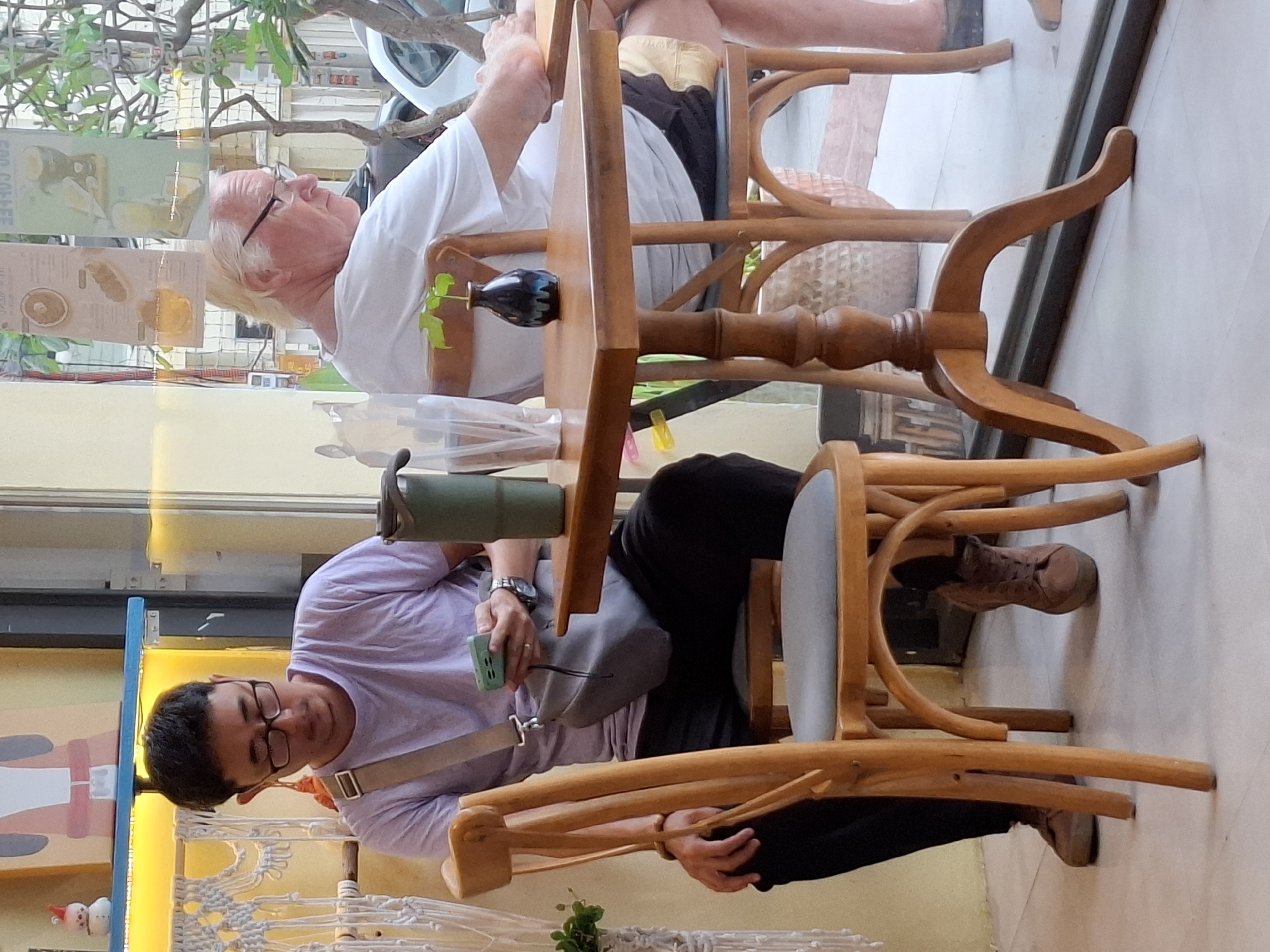 Two men are seated at tables inside a cafe in Nha Trang, Vietnam. The man on the left, wearing a lavender t-shirt, glasses, and black pants, is holding a green smartphone and looking down at it. A grey bag is slung across his shoulder. A green insulated tumbler sits on the table in front of him. He is seated in a wooden chair with a grey cushion.
The man on the right, with blond hair and glasses, wears a white t-shirt and dark shorts. He is looking to his right, away from the camera. A small dark vase with a single green leaf is on the table in front of him. He is seated in a wooden chair with a grey cushion, partially obscured by the table.
The cafe has a brightly lit interior with yellow and white decor visible in the background. The scene appears to be viewed through a large window, with reflections of trees and vehicles visible on the exterior. The tables and chairs are made of wood.