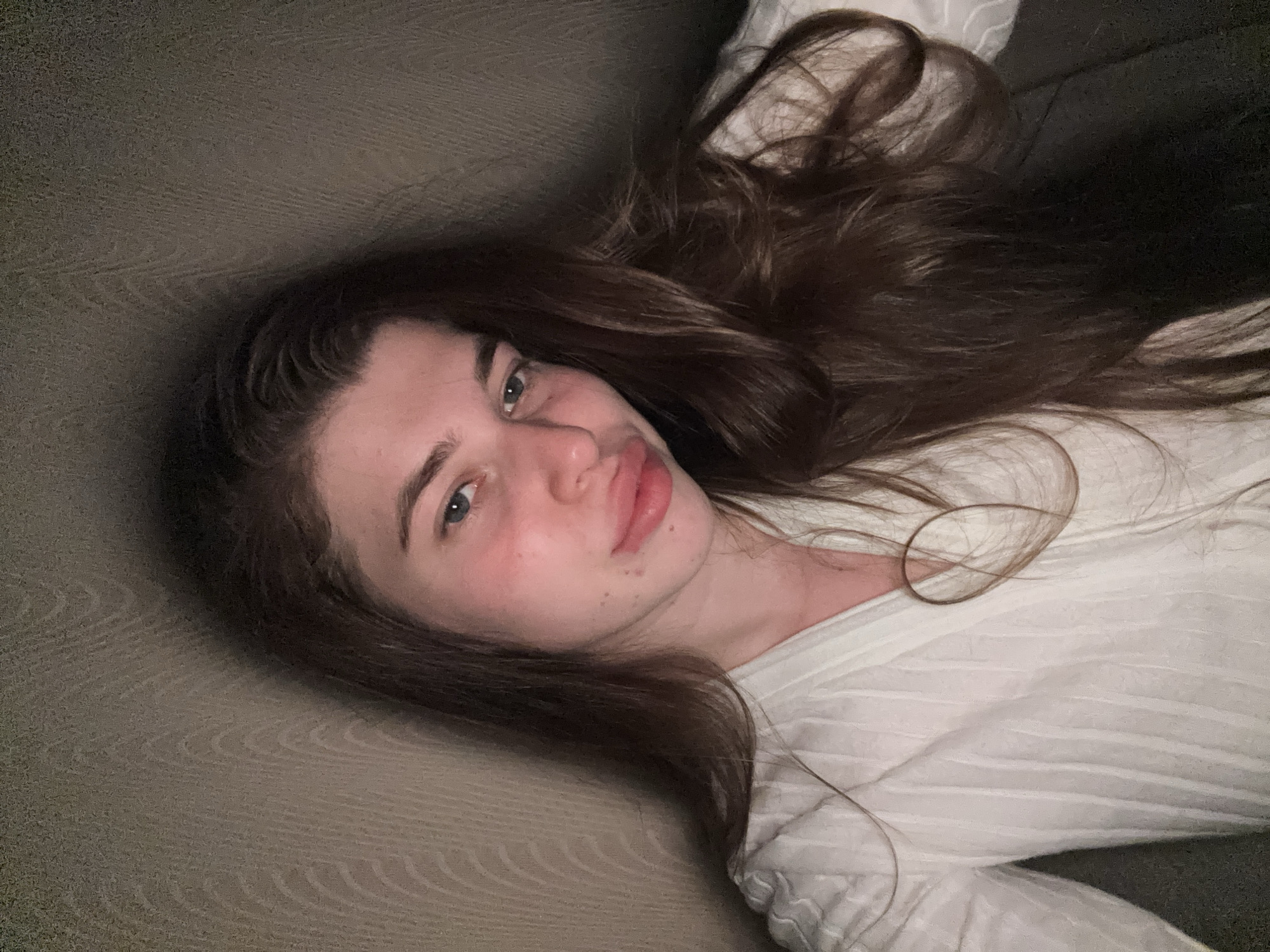 A woman lies on a neutral-colored sofa. She has long, brown hair spread around her head. She is wearing a white, textured long-sleeved shirt. Her eyes are open and she is looking at the camera. The background is slightly blurred. The setting appears to be indoors, likely a home. There is no information in the image to suggest a location in the city of Мещёры, Russia.