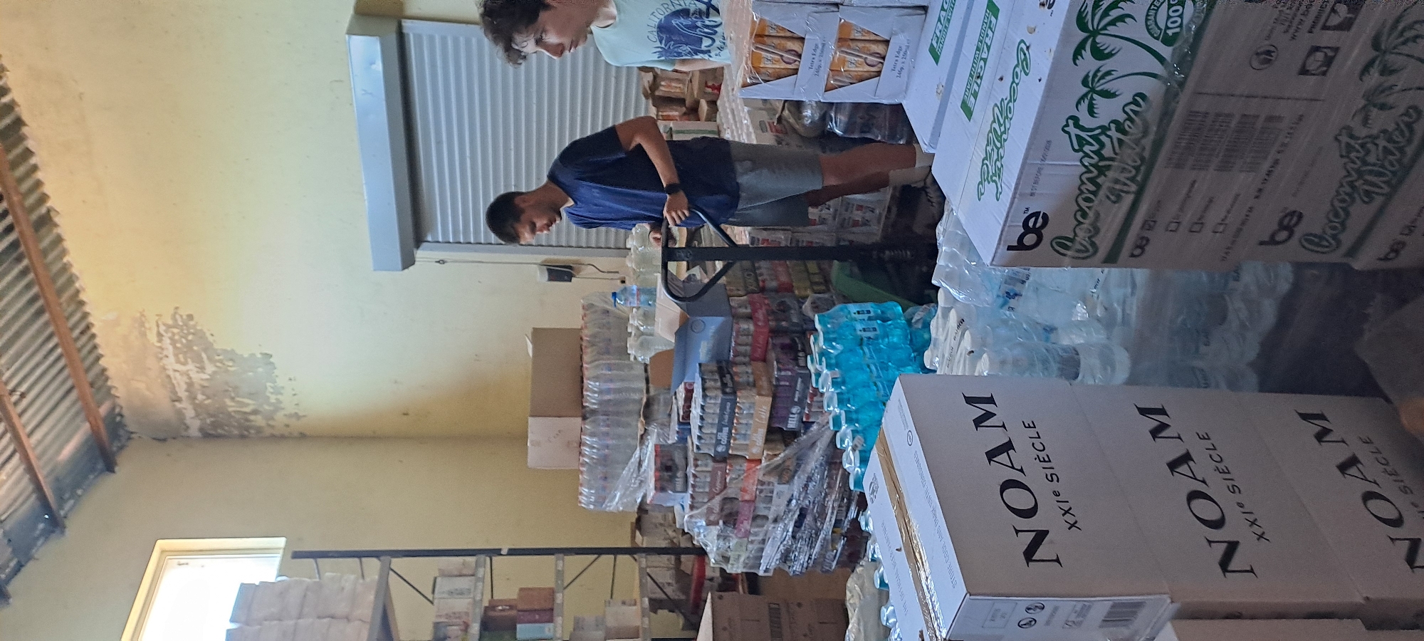 The image shows the interior of a storage facility in Velika, Bulgaria, filled with stacks of bottled water and other beverages. Two young men are present, appearing to be organizing or preparing the goods. One man, wearing a dark blue t-shirt and grey shorts, is operating a pallet jack, which is positioned near a pallet of boxed drinks. He is looking down at the pallet jack. To his right, another young man, wearing a light blue t-shirt with a graphic print, is looking towards the right side of the frame. The environment appears to be a warehouse or storage unit, with corrugated metal roofing visible overhead and pale yellow walls. Shelving units with boxes are present on the left side of the image, and large stacks of boxed and bottled products dominate the central and right portions of the scene. The overall impression is one of a logistics or distribution point for beverages.