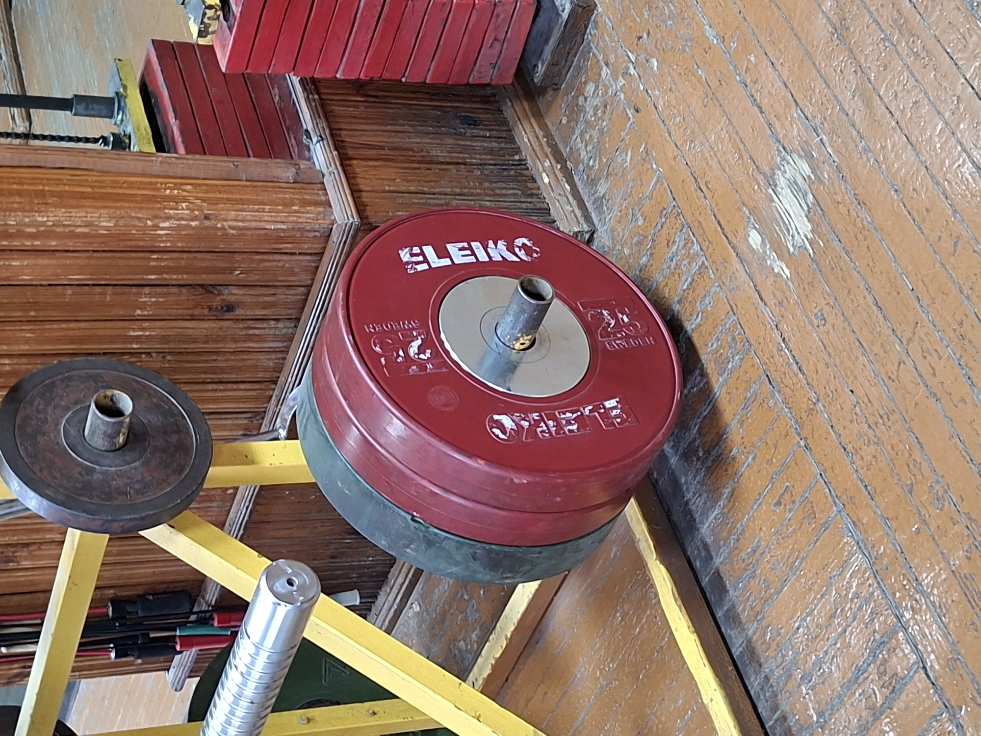 The image shows a barbell with red weight plates, manufactured by Eleiko and weighing 25 kg each, in what appears to be a gym or a weightlifting facility. The plates are stacked on a yellow metal rack, which also supports a smaller, darker weight plate on top. In the background, there are red weight stacks and wooden paneling. The floor is made of worn wooden planks. The scene is static and does not depict any individuals or actions. There is no identifiable information in the image to suggest a specific location within Kazan, Russia.