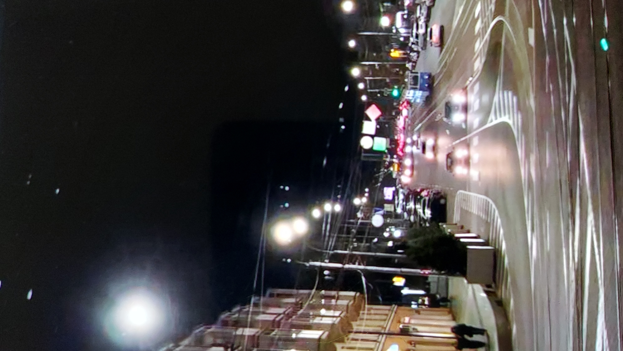 An outdoor, medium-shot, nighttime image depicts a city street with traffic and pedestrian activity. On the left, a multi-story building with arched windows lines the street. Two pedestrians are walking on the sidewalk in front of the building. The street is illuminated by streetlights and the headlights of passing cars. A tram is visible in the distance, and several cars are driving on the road. The sky above is dark and appears to have some celestial bodies or atmospheric phenomena, with a bright, diffused light source in the upper left quadrant. The road surface shows painted lane lines and what appear to be tram tracks.