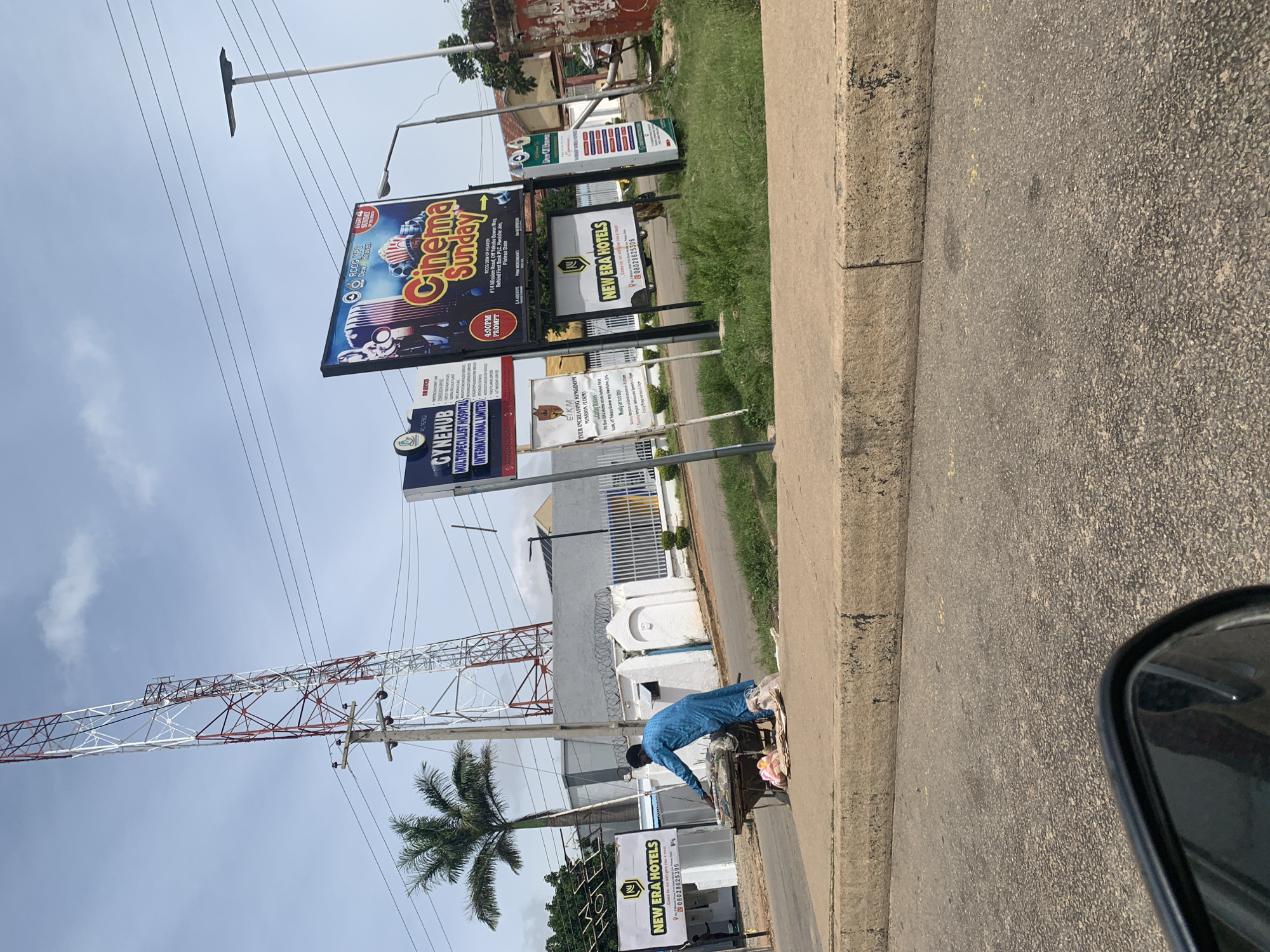 The image depicts a street scene in Jos, Nigeria, characterized by a tall telecommunications tower in the background. In the foreground, a person wearing a blue kaftan is pushing a cart laden with what appears to be assorted goods, possibly for sale or transport. The cart is positioned on the side of the road, next to a concrete curb. Several billboards are visible, advertising "New Era Hotels," "Gynehub Multispecialist Hospital," and a "Cinema Sunday" event. The environment suggests an urban or semi-urban setting, with roads, sidewalks, and buildings. The sky is partly cloudy, indicating daytime. The perspective appears to be from a vehicle, as a portion of a side mirror is visible in the lower left corner.