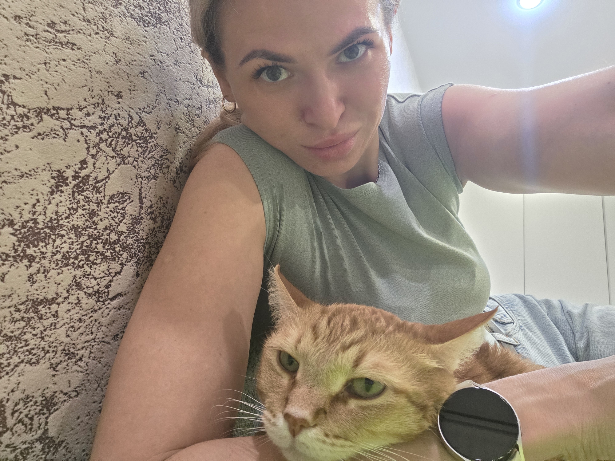 A woman with blonde hair and green eyes is holding a ginger cat. The woman is wearing a green t-shirt and a denim jacket. She is also wearing a smartwatch. The background is a wall with a patterned design and a white wall. The location is in the city of Chaikovsky, Russia.