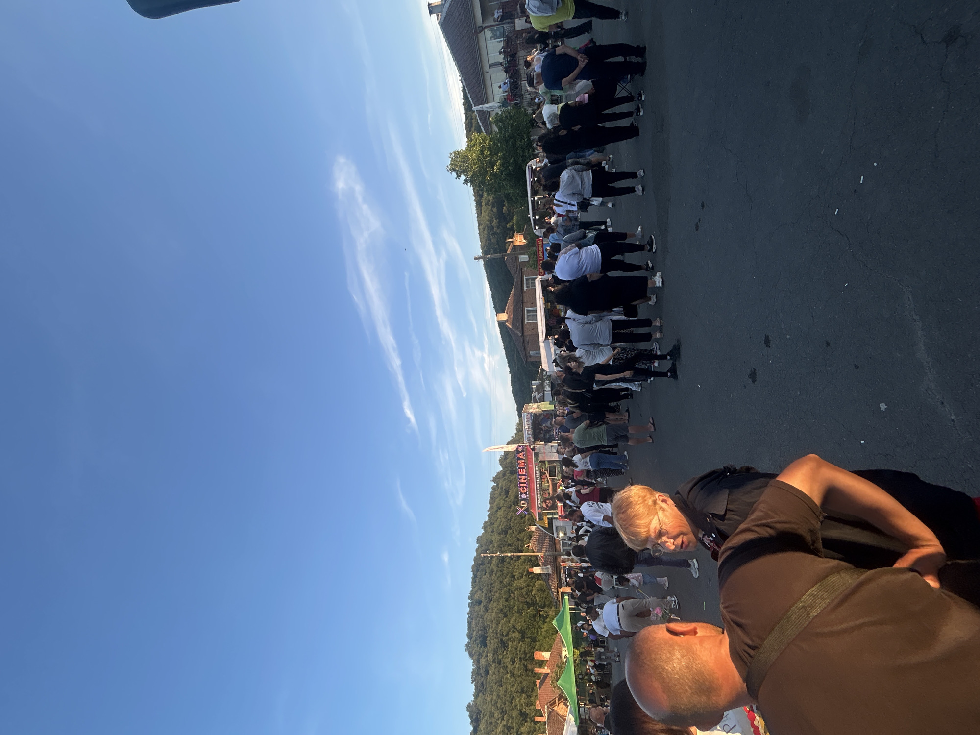 In the city of Gabar, Bulgaria, a public gathering takes place on a paved street. A clear blue sky with wispy clouds stretches above, casting a warm, late afternoon light. The scene is populated by a diverse crowd of people, many of whom are casually dressed, suggesting an outdoor event or festival.
Prominently featured is a large banner with the words "3D CINEMAS" and "ATTRACTION VIRTUAL REALITY," indicating a form of entertainment is present. Tents, possibly for vendors or attractions, are set up along one side of the street, with people milling around them. In the background, traditional-looking buildings with red-tiled roofs are visible, nestled against a backdrop of lush green hills.
Two individuals in the foreground are engaged in conversation. One man, viewed from the back, wears a dark t-shirt and has a bag slung across his body. He appears to be gesturing with his right hand. Facing him is a woman with short, auburn hair and glasses, wearing a dark jacket. Their interaction suggests a casual exchange within the bustling environment. The overall impression is of a lively community event unfolding in a scenic, rural Bulgarian setting.