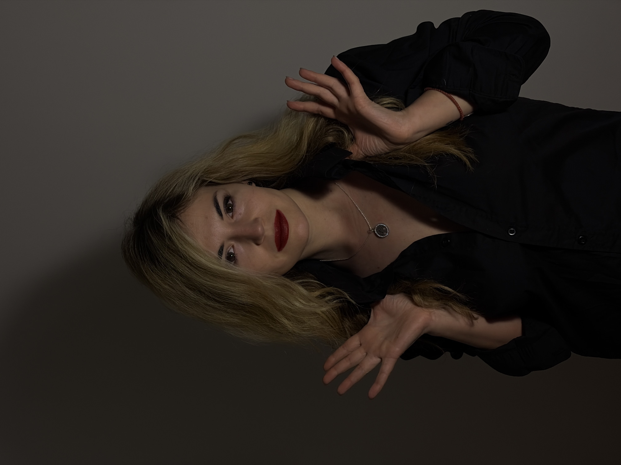 A woman with blonde hair and red lipstick is lying down, wearing a black shirt and a silver necklace. Her hands are raised with her fingers spread. The background is a plain gray. No information suggests the location is in Budva, Montenegro.