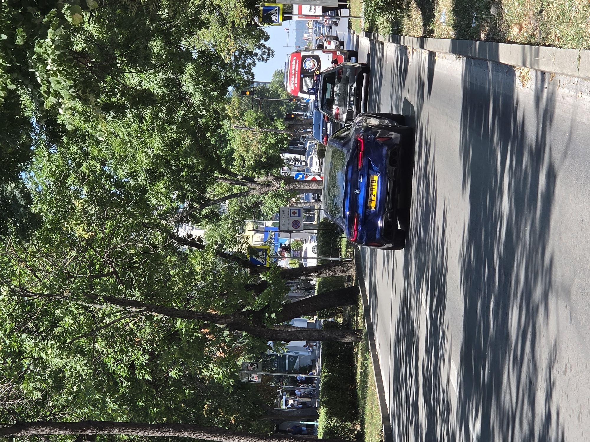 This image depicts a street scene in Varna, Bulgaria, likely during daylight hours given the strong shadows cast by trees. The perspective is from a vehicle driving on a paved road.
In the foreground, a dark blue BMW M8 coupe is prominent, seen from its rear. It has a yellow license plate with the inscription "Y2 OTO". The car is moving forward.
Several other vehicles are visible on the road, including a white van with red and white lettering that appears to be an ambulance or medical transport, a dark colored Volkswagen Golf, and other cars further down the road. Traffic lights are present, one of which is illuminated yellow.
The street is lined with mature trees, their dense foliage creating a dappled pattern of light and shadow on the road surface. Sidewalks are visible along the side of the road, with some pedestrians walking on the left side. Various street signs and traffic signs are present, including pedestrian crossing signs and what appear to be directional or informational signs in Bulgarian. The environment suggests an urban setting with a park-like ambiance due to the abundant trees.