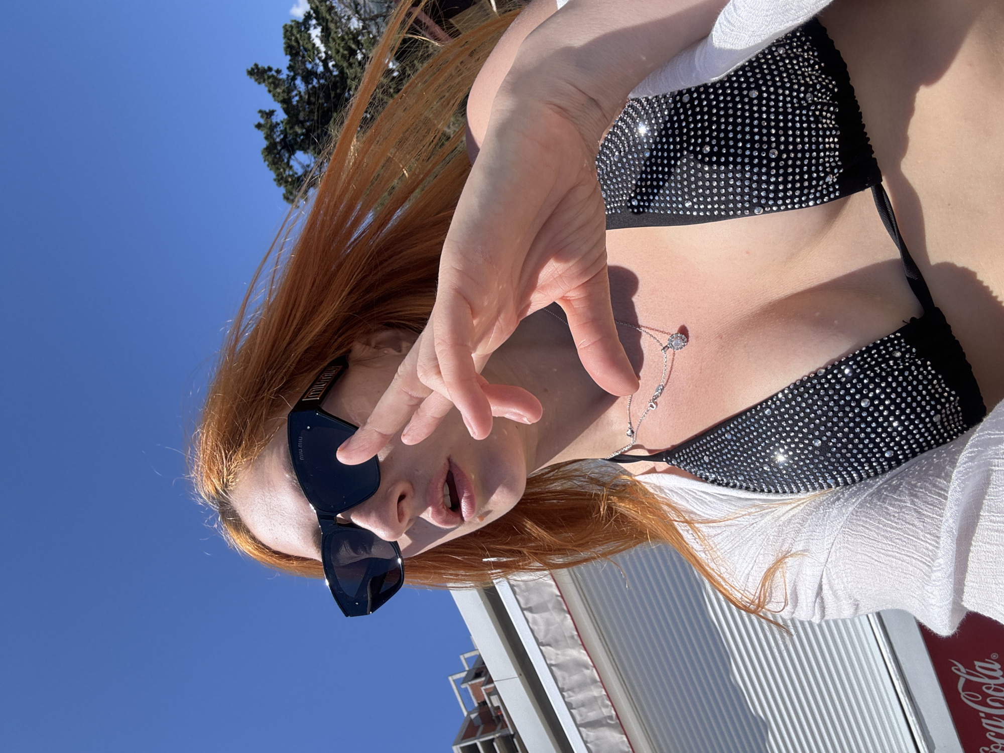 A vibrant, low-angle portrait captures a person, likely female, with long, flowing reddish-brown hair under a clear blue sky. She is wearing black sunglasses, with the brand "Miu Miu" visible on the arm, and a black bikini top heavily embellished with small, sparkling rhinestones. Over the bikini top, she wears a light-colored, possibly white, textured cover-up or shirt. A delicate silver necklace with a circular, sparkling pendant adorns her neck.
Her right hand is raised, partially obscuring her face, with her fingers spread slightly and her mouth open. The strong sunlight casts shadows and highlights the glitter on her top. In the background, a deep blue, cloudless sky dominates, with the dark green branches of a tree visible in the upper left corner. Towards the bottom right, parts of a modern building with horizontal siding are visible, along with a prominent red sign bearing the white "Coca-Cola" logo. The bright lighting suggests it is daytime, under sunny weather. The scene evokes a relaxed, outdoor atmosphere, consistent with a leisure setting in Budva, Montenegro.