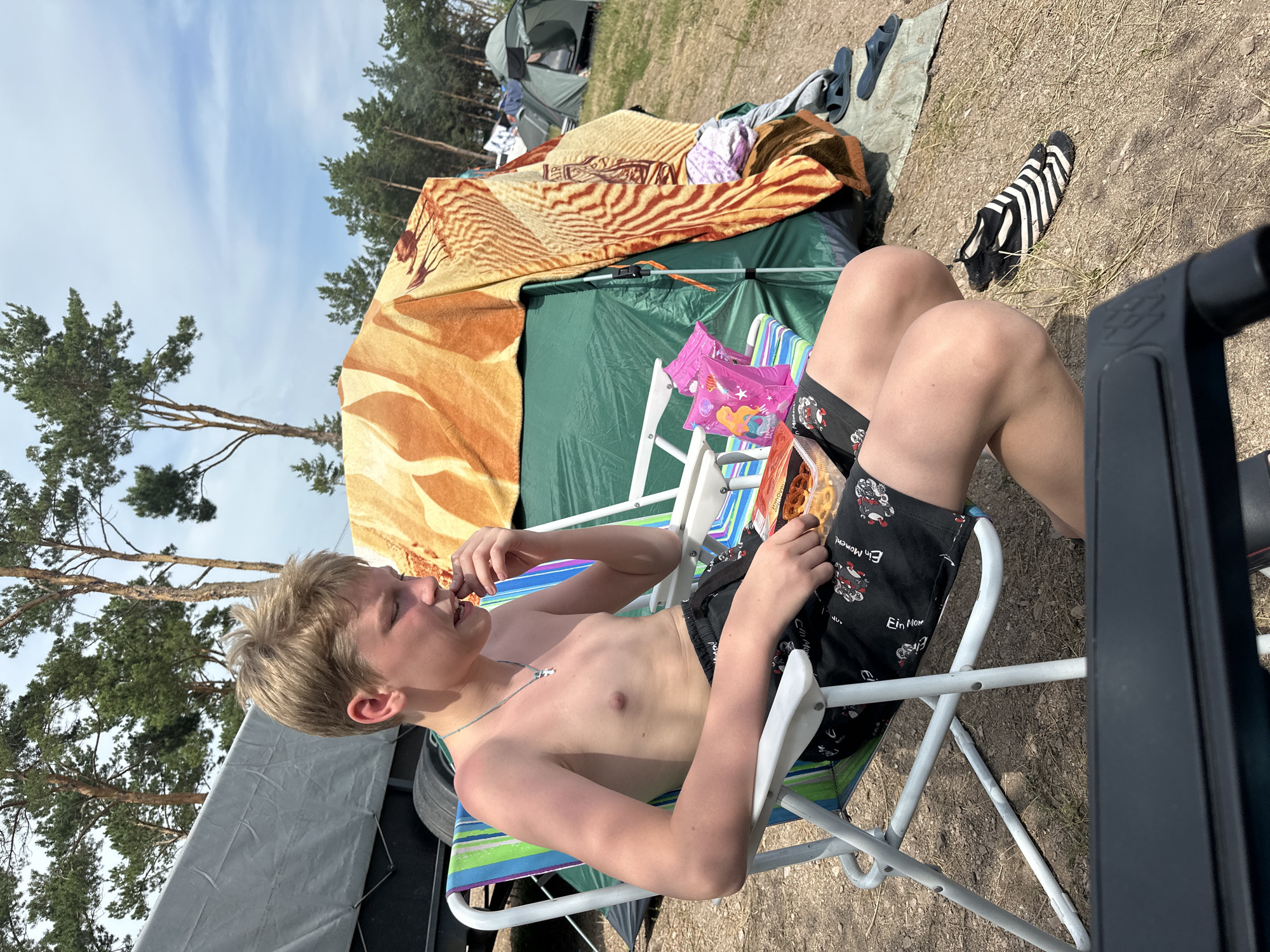 A young boy with blond hair is sitting in a striped camping chair, facing right. He is shirtless and wearing black shorts with a pattern of motorcycles and the German phrase "Ein Moment". He is eating from a bag of chips held in his right hand, with his left hand near his mouth. The boy has a relaxed expression.
The scene is a campsite with tents and trees in the background. A green tent is visible behind the boy, partially covered by a patterned blanket with a tiger-like stripe design. There are other camping items scattered around, including a pair of black and white striped socks on the ground and some dark blue flip-flops. The ground is covered in dirt and dry grass.
The sky is blue with some wispy clouds, suggesting a sunny day. The lighting indicates it is daytime, likely afternoon. The presence of camping gear and the outdoor setting place the scene at a campsite. The overall atmosphere appears to be casual and relaxed, consistent with a camping trip. The text "Ein Moment" is visible on the boy's shorts.