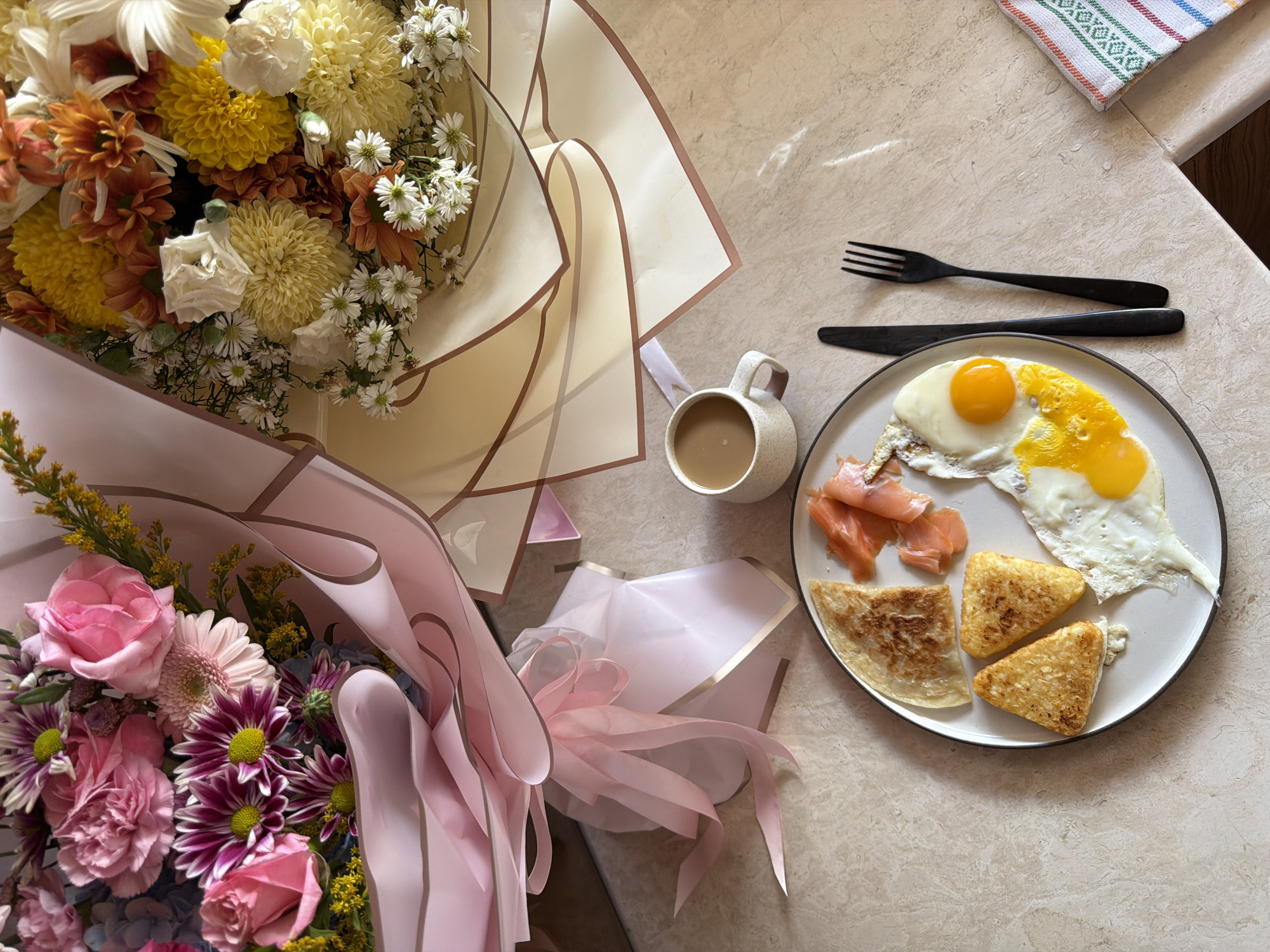 On a light, patterned surface are two bouquets of flowers, a plate of food, a mug, and silverware. One bouquet has yellow, orange, and white flowers with a cream-colored wrapping, while the other has pink and purple flowers with a pink wrapping. The plate contains two fried eggs, smoked salmon, and hash browns. The mug is filled with a light brown liquid. A black fork and knife sit next to the plate. The surface is light in color with a subtle pattern. A corner of a patterned towel is visible in the upper right corner of the frame. There is no indication that the image was taken in Mengwi, Indonesia.