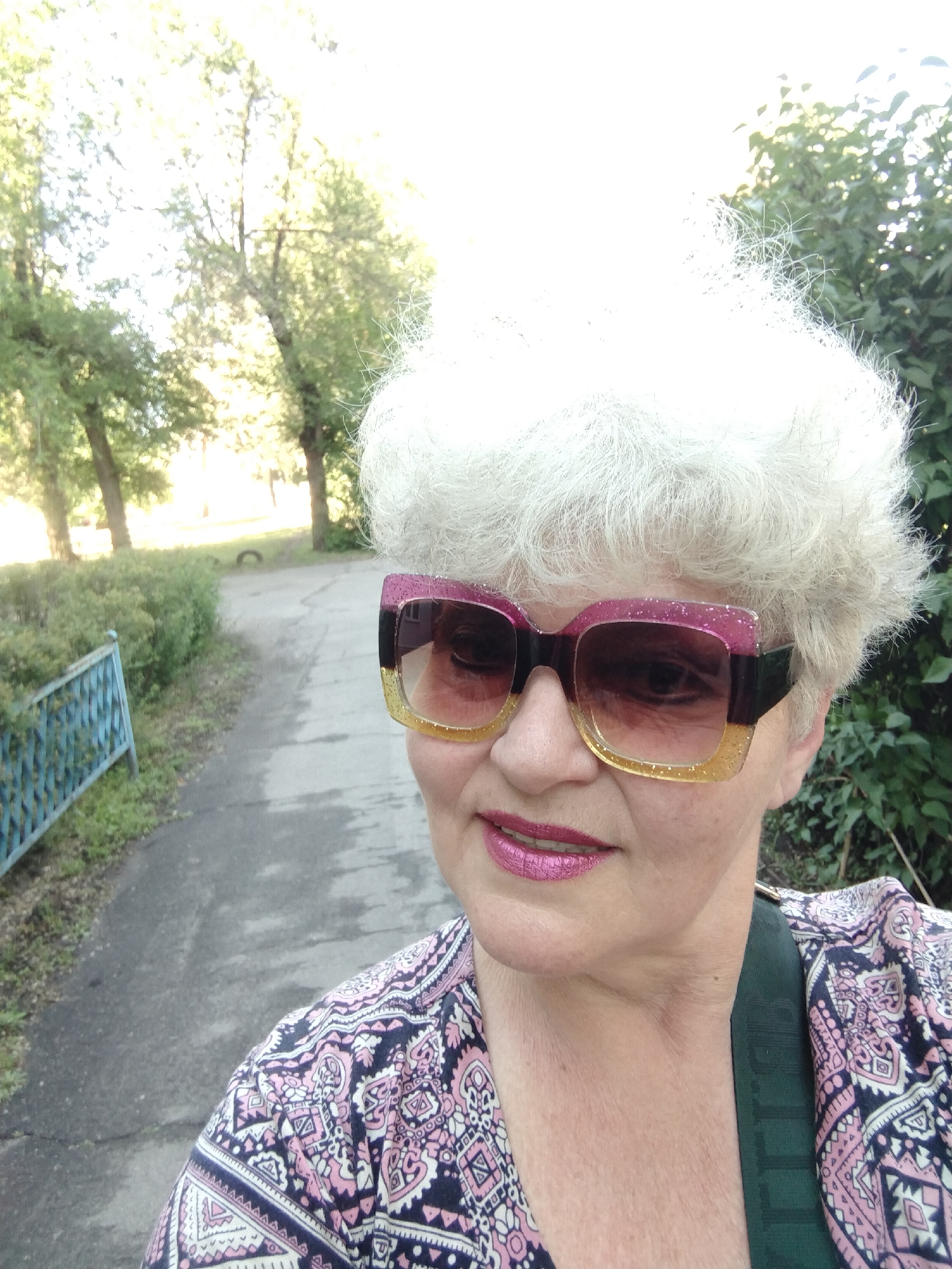 A woman is captured in a selfie outdoors in Horishni Plavni, Ukraine, on a bright, sunny day. She has short, curly, white or light grey hair. Her face is partly obscured by large, square-framed sunglasses that feature a distinctive two-toned glitter design: pink on the upper half and gold on the lower half, with dark tinted lenses. She is wearing bright pink lipstick. Her attire includes a blouse with a complex pattern in shades of pink, white, and a dark color, possibly navy blue or black. A dark green strap, likely from a bag, is visible across her right shoulder.
The background reveals a paved or asphalt path extending behind her. To her left, a low blue metal fence runs parallel to the path, backed by green bushes. Further back, several trees with green foliage are present, illuminated brightly by the sun, suggesting either morning or late afternoon. The path appears to recede into the distance, hinting at a park, pathway, or similar outdoor setting. The woman displays a relaxed expression with a subtle, pleasant smile. No visible text is present in the image.