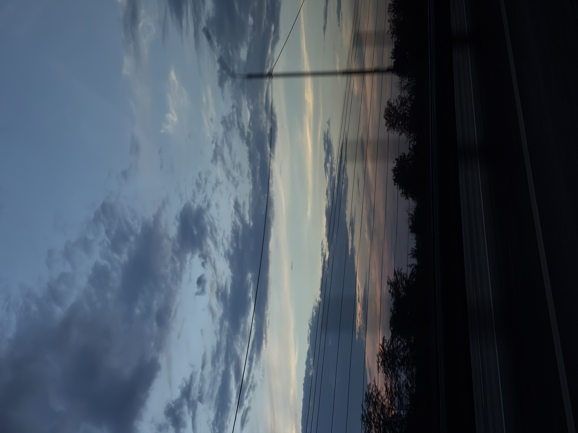 The image depicts a twilight sky with scattered clouds, transitioning from deep blue at the top to lighter hues near the horizon. Several power lines are visible horizontally across the frame, obscuring a silhouette of trees and vegetation along the bottom. A dark, possibly asphalt, road with white lines runs along the foreground. The overall scene appears to be captured from a moving vehicle, suggesting a view from a road or highway. The specific location in Lazurny, Russia, is not discernible from the visual information provided.