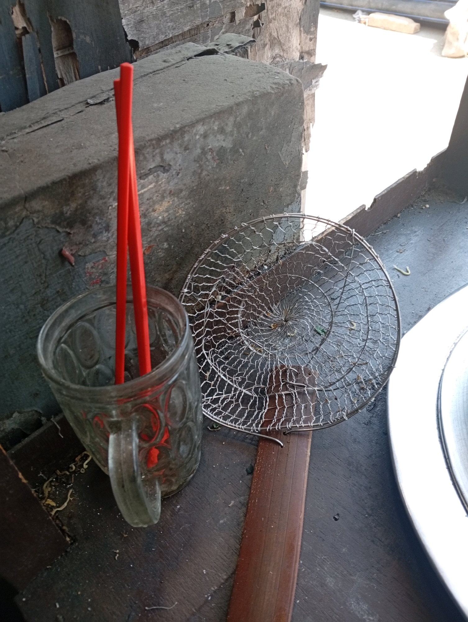 The image depicts a close-up, outdoor scene, likely a food stall or cooking area in East Jakarta, Indonesia, with a focus on cooking utensils. In the foreground, a clear glass mug with a handle contains two bright red chopsticks. The mug appears to be resting on a dark, weathered wooden surface. To the right of the mug is a round metal strainer with a wooden handle. The strainer is positioned diagonally across the frame and shows signs of use, with small food particles visible within its mesh. The background consists of a rough, grey concrete structure and a wooden plank, suggesting an outdoor, possibly rustic, setting. The lighting indicates it is daytime, with bright light sources illuminating parts of the scene.