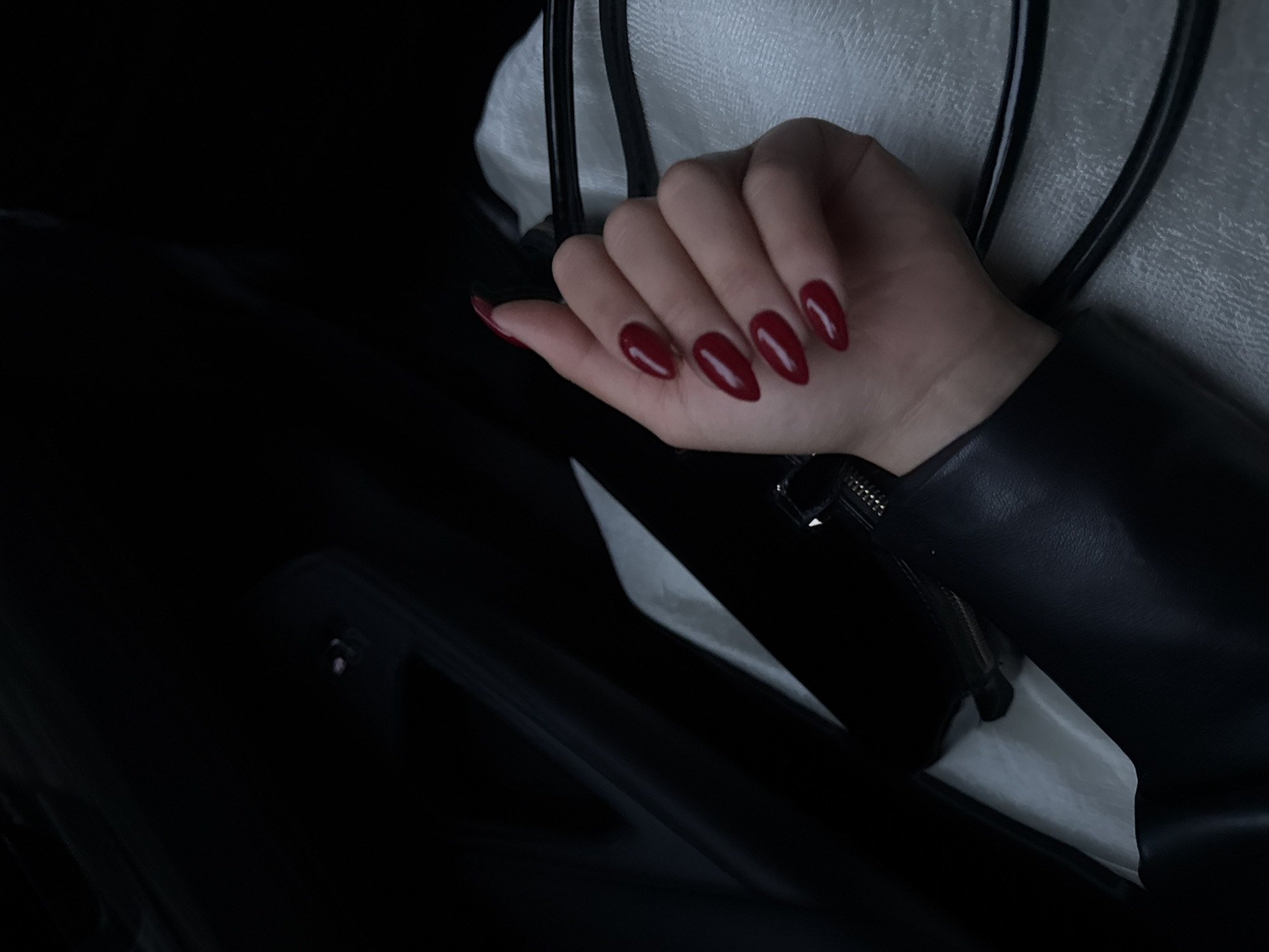 A hand with red nail polish rests against the inner black leather of what appears to be a vehicle door. The hand, positioned in the foreground, features almond-shaped nails reflecting light. The background contains a white, perhaps fabric-covered, object and more black leather material. No indication of Yerevan or Armenia is visible. The scene conveys a sense of confinement and sleekness.