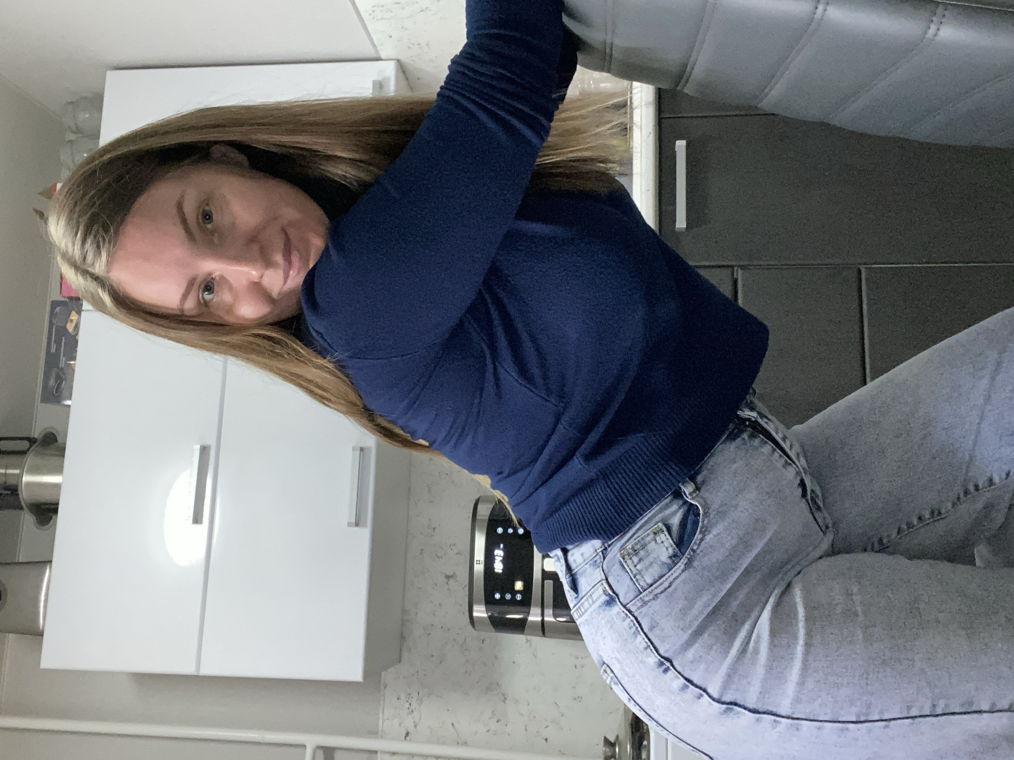 This image captures a young woman posing in a modern kitchen setting in Noviy, Russia. The photograph is rotated 90 degrees counter-clockwise. The main subject is a woman with long, straight brown hair and light eyes, looking directly at the camera with a gentle, slightly smiling expression. She is dressed casually in a navy blue long-sleeved top and light-wash blue jeans.
The background details a contemporary kitchen. On the left, there are white upper cabinets featuring sleek silver bar handles. On the right, darker grey lower cabinets with similar silver handles are visible. A light-colored countertop, possibly with a speckled or marbled pattern, runs across the kitchen. A prominent black and silver kitchen appliance, likely an air fryer, sits on the countertop. Its digital display clearly shows the time "10:43". Part of a padded, grey chair or stool is visible in the upper right quadrant of the image. A metallic pot or pan is partially seen in the lower-left corner. The overall scene suggests a relaxed moment within a clean and modern home environment.