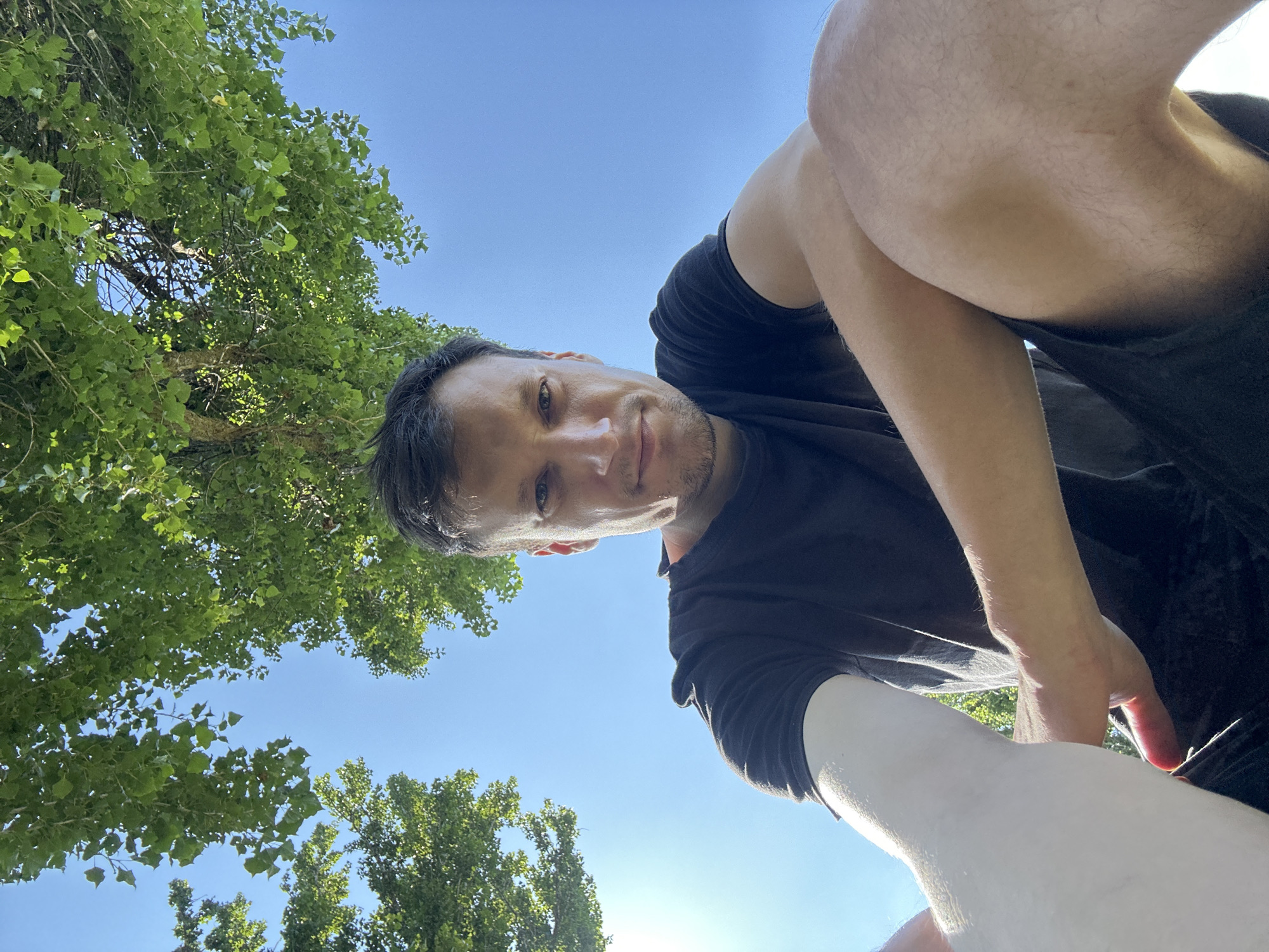 A man is captured in a low-angle, wide-shot selfie, taken outdoors under a bright blue sky. He is seated, with his legs crossed, in what appears to be a park or natural setting. Lush green foliage from a tree hangs overhead, providing a partial canopy. The man is wearing a dark t-shirt and dark shorts. The sun is shining brightly, casting a glow on his skin and parts of his arm, and the image appears to have been taken on a clear, sunny day. No specific landmarks identifying the city of Makiv, Ukraine, are visible.