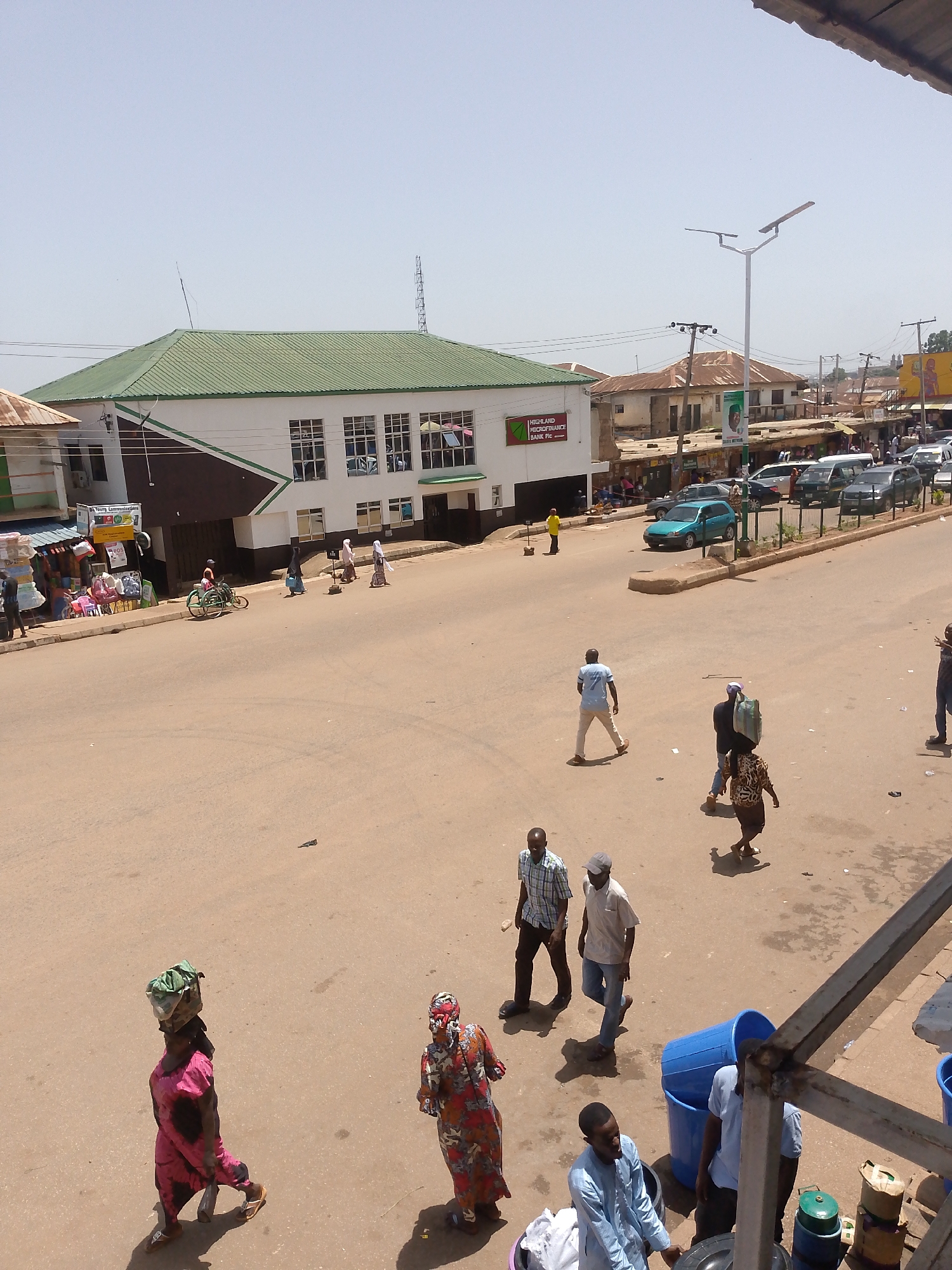 The image depicts a street scene in Bukuru, Nigeria, under a bright, sunny sky. The street is paved and dusty, with multiple people walking across it. Some individuals are carrying items on their heads. Vehicles, including cars and vans, are parked along the side of the road. Buildings line the street, including a white building with a green roof identified as "Highland Microfinance Bank Plc." There is a streetlight visible, and a telecommunications tower rises in the background. Various commercial activities appear to be taking place along the sidewalks, with vendors and displays of goods. Blue plastic containers are observed in the foreground.