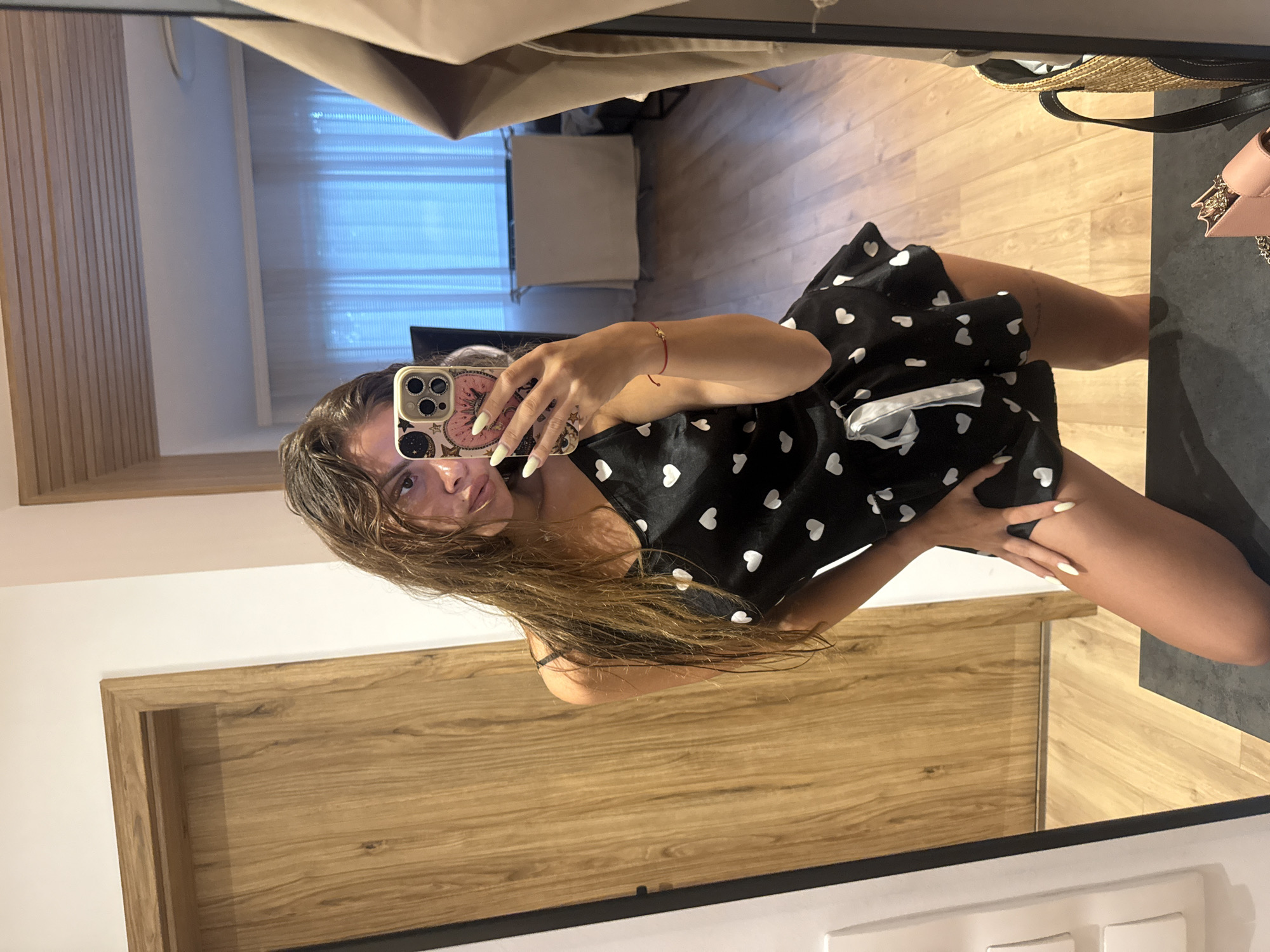 The image shows a young woman taking a mirror selfie in what appears to be a bedroom. She is wearing a black slip with white hearts and a white drawstring. Her hair is long and brown, and she is wearing light-colored nail polish. In her hand, she is holding a smartphone with a decorative case. The background shows a wooden door, a reflective surface that could be a window or another mirror, and a floor with a wooden pattern. There is also a glimpse of a tan coat hanging in the reflection. The overall environment suggests a domestic setting, likely a home. There are no specific indicators that place the scene in Plovdiv, Bulgaria, or any particular city.