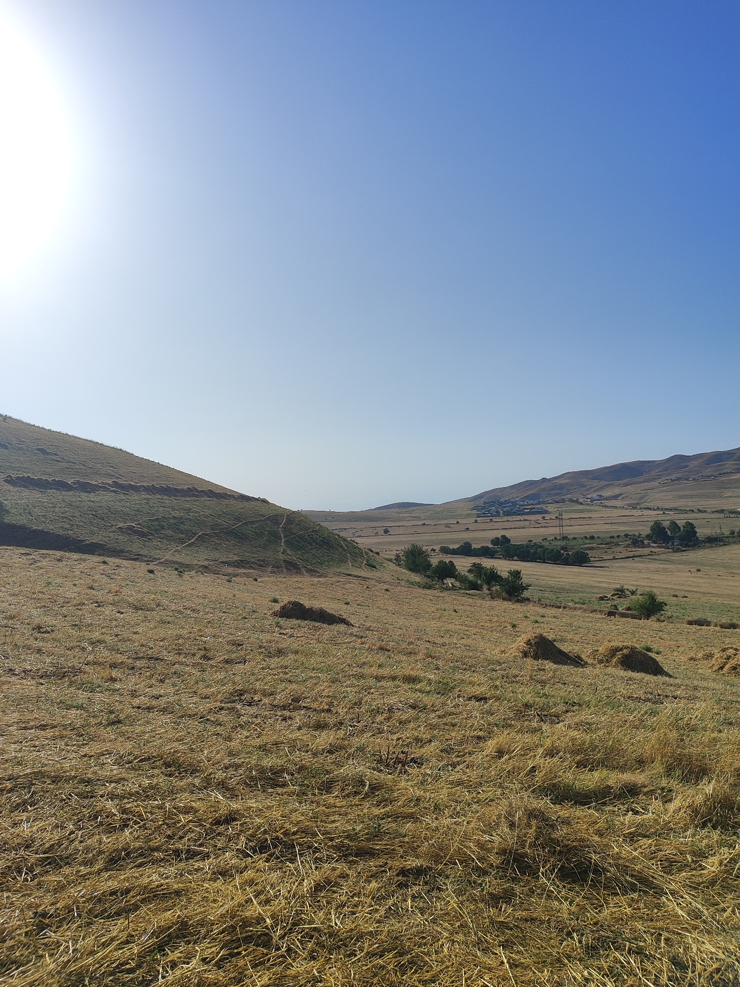 A wide-angle, eye-level, outdoor, daylight, horizontal shot captures a rural landscape under a clear, bright blue sky. The sun shines brightly from the upper left corner, casting a soft light over the scene. In the foreground, a field of dry, golden-brown grass covers the ground, with several piles of harvested hay scattered across it. To the left, a grassy hillside slopes upwards, marked with faint trails. In the middle ground and background, the landscape transitions to a valley with more flat, dry fields, dotted with trees and a small village in the distance. Rolling hills form the backdrop, their slopes textured with vegetation and some scattered buildings. A power line tower is visible in the valley. The overall impression is one of a dry, agricultural, and somewhat sparse countryside on a sunny day.