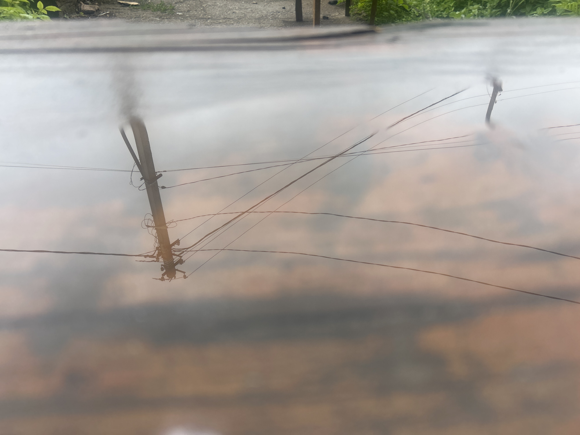 The image is a reflection of a utility pole and power lines in a puddle of water. The water's surface is calm, creating a clear, albeit upside-down, reflection of the objects above. The utility pole is made of concrete and has several wires attached to it. The wires stretch across the frame, some taut and others slack. The reflection shows a blurred background of what appears to be a street or pathway with some greenery. The color palette is muted, dominated by grays and browns, with hints of green from the foliage. The overall impression is one of a quiet, perhaps overcast day. The location is the city of Ignatovo, Russia.