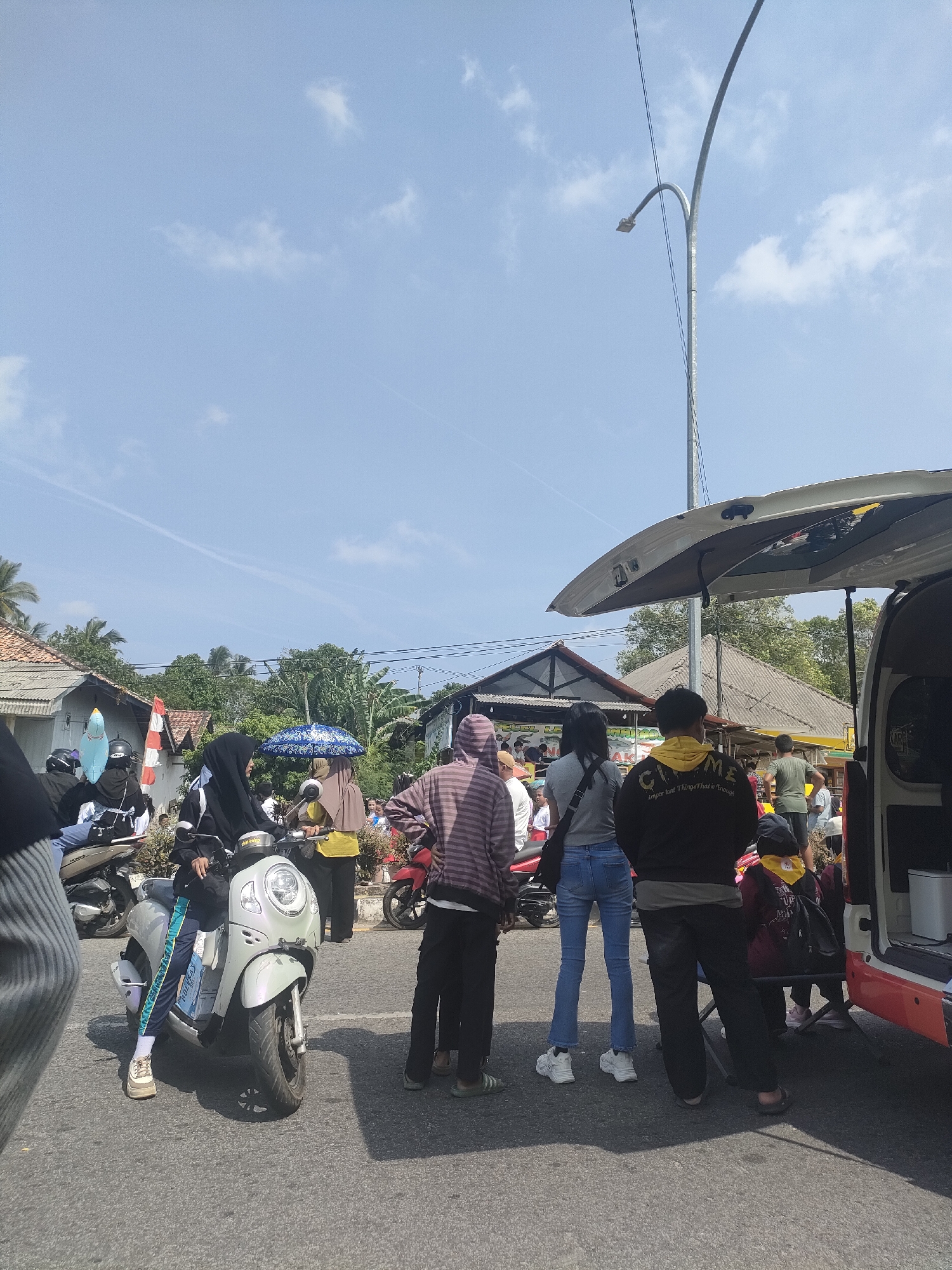 The image depicts an outdoor scene in Sungailiat, Indonesia, on a bright, sunny day with a clear blue sky and scattered clouds. The foreground is dominated by a paved road. On the left side, a light-colored scooter is parked, with a person seated on it wearing a black hijab, patterned leggings, and white sneakers. To the right of the scooter, a group of people are standing, facing away from the camera. Among them, a person in a striped hoodie and dark pants is prominent, as is a person in a black hooded sweatshirt with yellow lettering that reads "CHAMELEON important things that is enough" and dark pants. Next to them, a person in a grey t-shirt and blue jeans is visible. In the background, several buildings with tiled roofs are visible, surrounded by lush green trees. A white van with its rear door open is parked on the right side of the frame. A street lamp curves over the scene. Several other individuals and a red motorcycle are also present in the background, suggesting a public gathering or street scene.