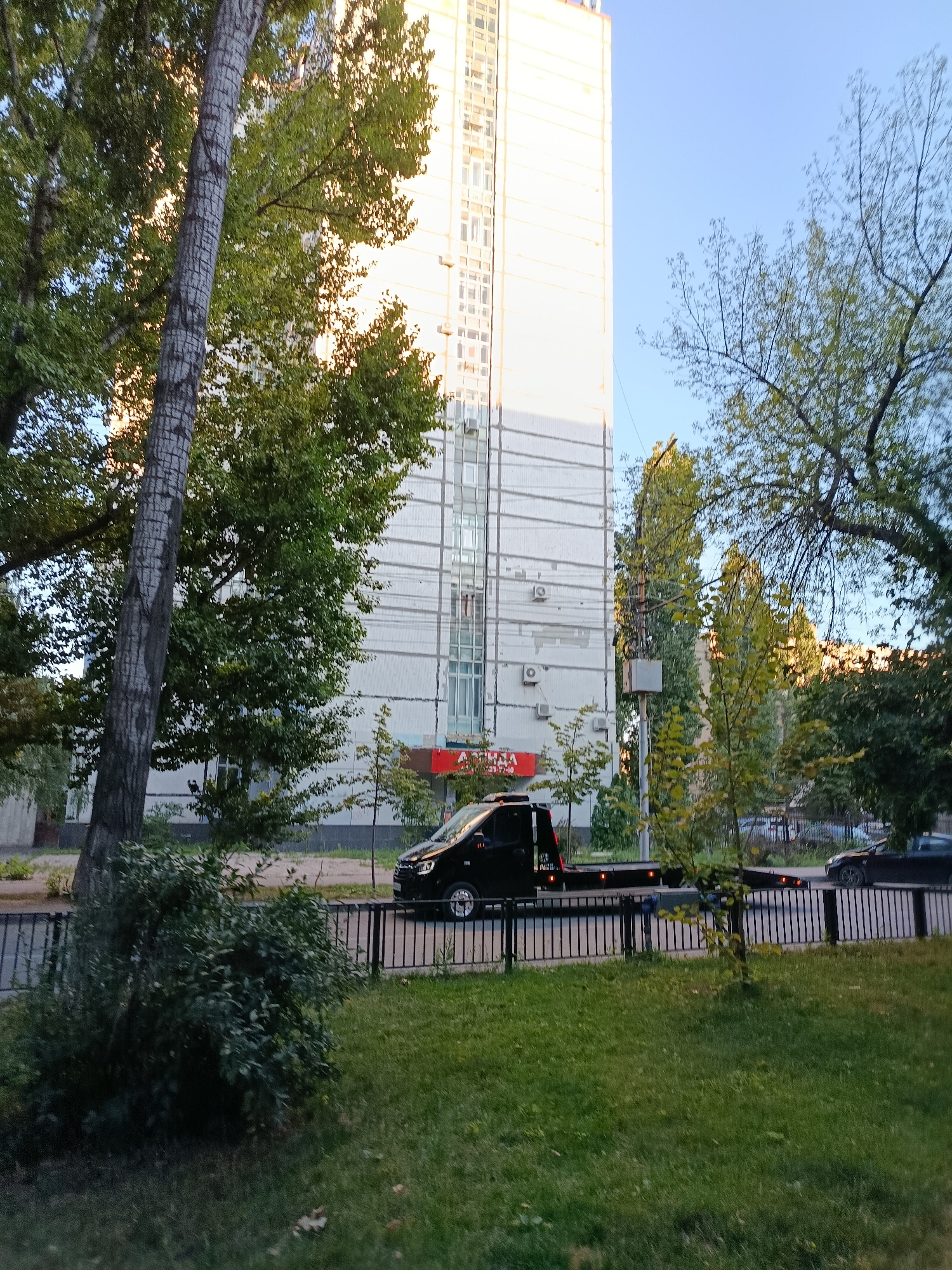A flatbed tow truck is parked on the side of a street in Saratov, Russia, with a tall apartment building in the background. The tow truck is black with amber lights on the rear. A black car is parked further down the street. Trees and grass are visible in the foreground. The sky is clear and blue.