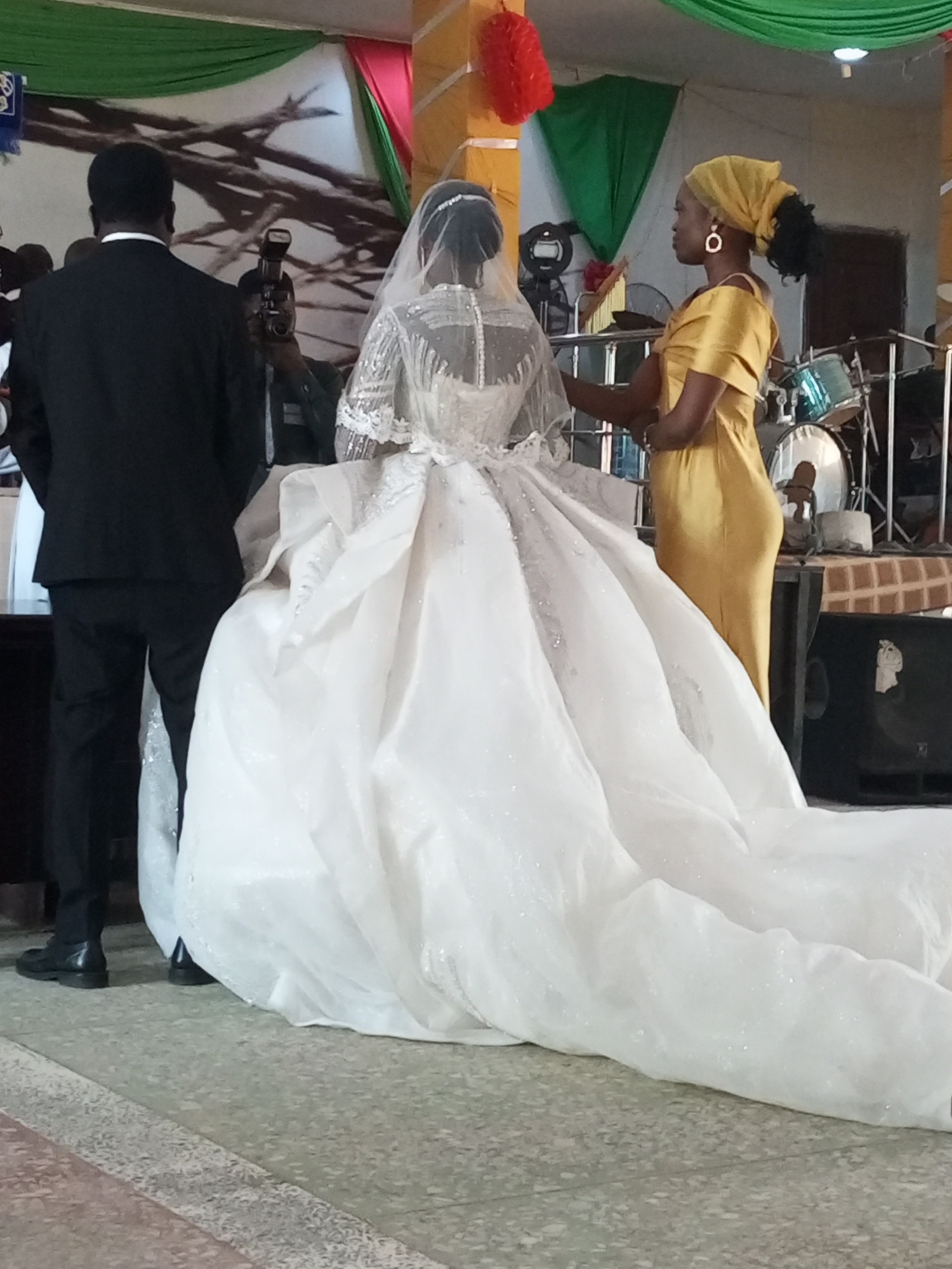 The image captures a wedding scene, likely indoors, given the presence of decorations and what appears to be a stage with musical instruments like drums. The focal point is the bride, wearing a large, elaborate white wedding gown with a long train. The groom stands beside her in a black suit. A woman in a yellow dress and matching head wrap is positioned near the bride, possibly assisting with the dress or providing support. A photographer with a camera and flash is visible in the background, capturing the moment. The scene appears to be taking place in the city of Unknown, Unknown.