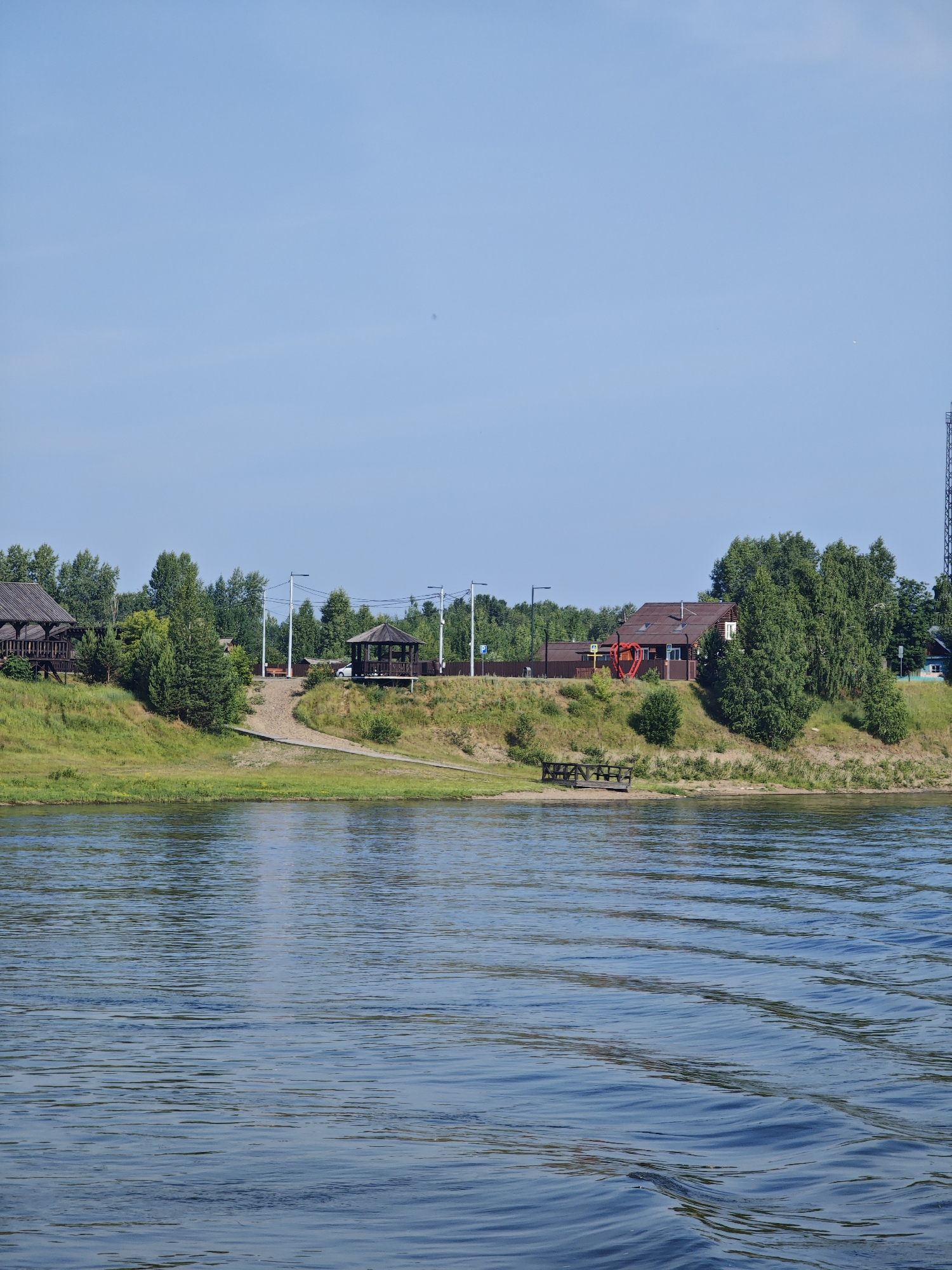 The image depicts a scenic view of a riverbank on a clear day. In the foreground, the blue water of the river displays gentle ripples, reflecting the sky and the surrounding greenery. The middle ground features a grassy embankment rising to a sandy area with a wooden path leading up to it. Several structures are visible on the embankment, including two wooden gazebos, one with a dark roof and lattice sides, and another with a lighter brown roof and open sides. A wooden house with a reddish-brown roof and white trim stands on the higher part of the embankment, adorned with a large red heart-shaped sculpture in front. Various trees, primarily deciduous and coniferous, are scattered along the embankment, creating a lush green backdrop. In the distance, a clear blue sky stretches across the horizon, with a few faint clouds. A tall radio tower is visible on the far right side of the image. The scene appears to be a tranquil riverside resort or recreational area, likely located in or near Galanino, Russia, given the context. There are no people visible in the image.