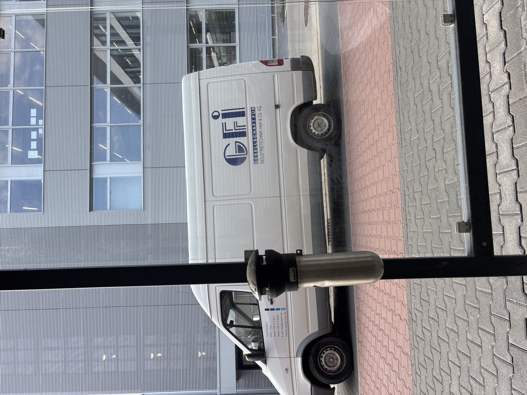 The image shows a white van parked in front of a modern building with horizontal metal siding and large windows. The van has a logo and text printed on its side, which reads "GFI" in large blue letters, followed by "GESELLSCHAFT FÜR INDUSTRIETECHNIK mbH & Co. KG." Below this, there is more text with an address and contact information. The van is parked on a red brick paved area, which transitions to grey interlocking pavers. A black pole with a silver cylindrical object attached to it is in the foreground, partially obscuring the view of the van and the building. The scene appears to be outdoors, under daylight. The text on the van indicates the company's location as Industriestraße 2, 64653 Lorsch. While the image is captured in Mannheim, Germany, the specific location of the van in Mannheim is not evident from the provided visual information.