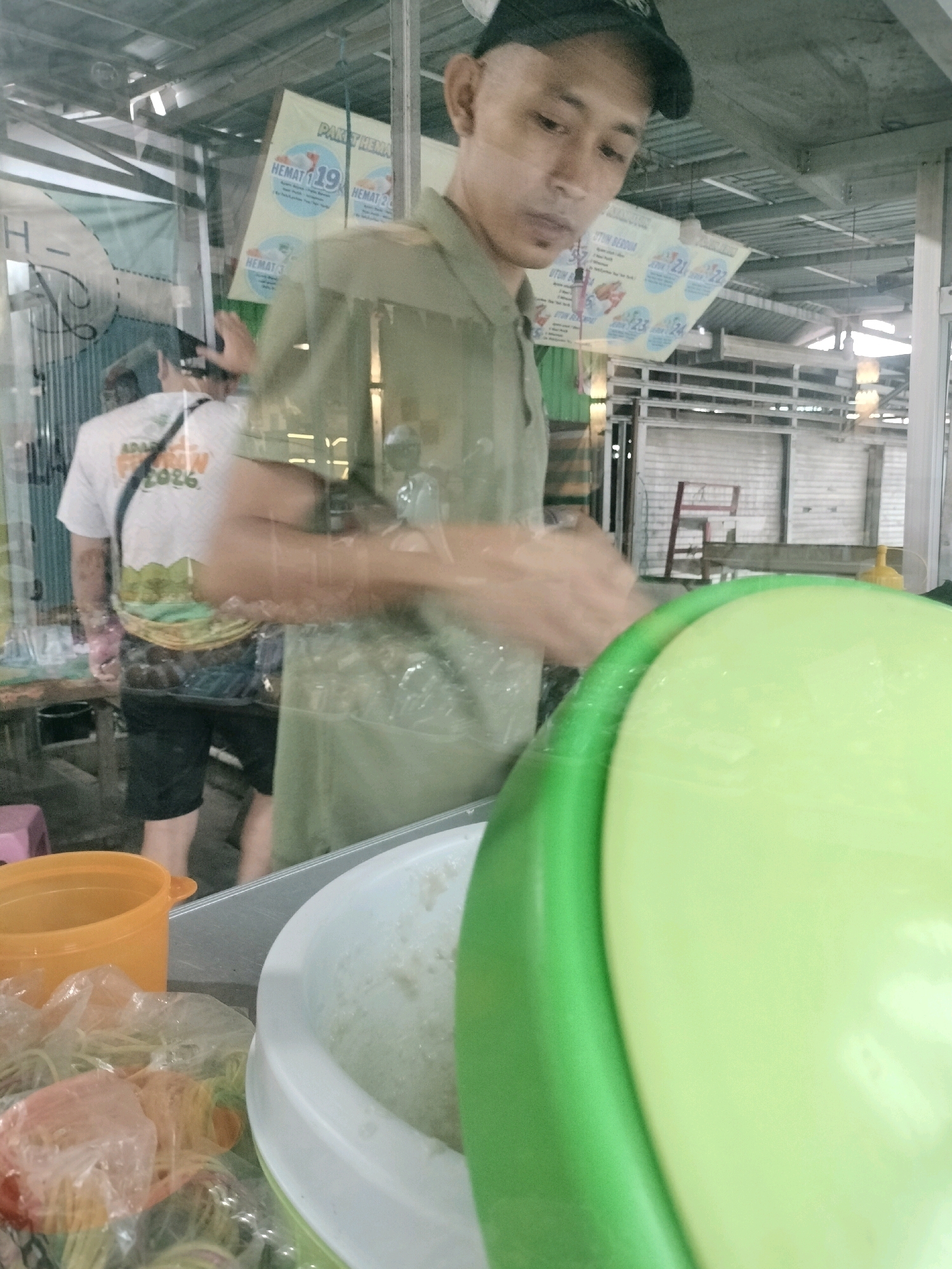 The image depicts a street vendor in Pembataan, Indonesia, preparing food. A man, wearing a dark baseball cap and a light green short-sleeved shirt, stands behind a food stall. His hands are blurred as he is in motion, likely serving or preparing food. In the foreground, a large, pale green bowl is visible, containing a light-colored mixture, possibly a dish being prepared. Next to it, an orange plastic container and several plastic-wrapped food items are present. Behind the man, another person wearing a white t-shirt with a graphic print and black shorts is visible, facing away from the camera. A banner with Indonesian text, including the word "HEMAT" (meaning "save" or "discount") and numbers like "19", "21", "23", and "24", is displayed in the background, suggesting a promotional event or sale. The overall environment appears to be an outdoor market setting, with glimpses of stalls, roofing, and corrugated metal structures. The lighting suggests it is daytime.