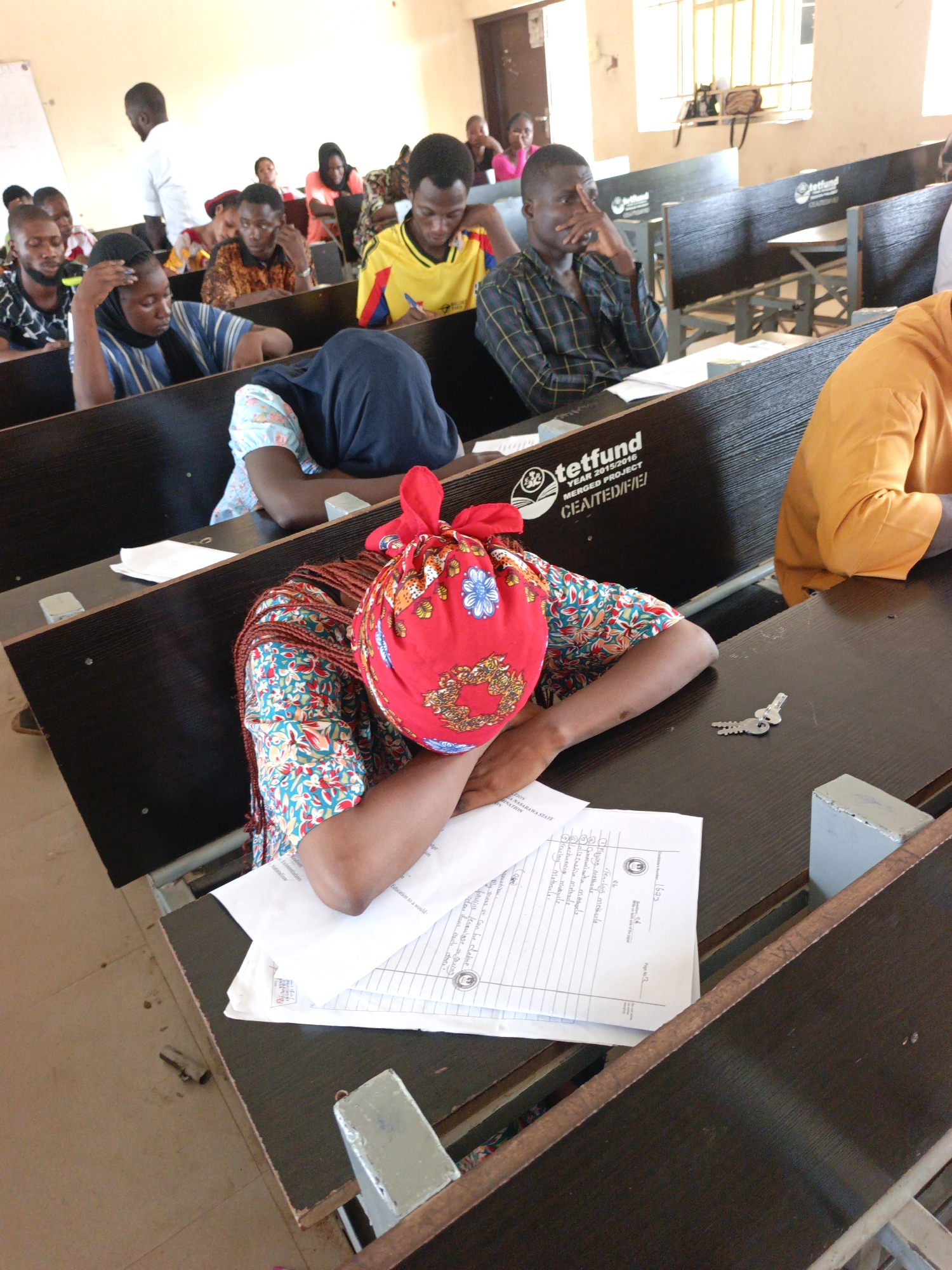 The image shows a classroom setting, potentially a university or college lecture hall, likely in Akwanga, Nigeria. Several students are seated at desks with “tetfund YEAR 2015/2016 MERGED PROJECT” printed on the side of the desks. A woman with a red headscarf has her head down on the desk, appearing to be sleeping or resting. There are papers and a set of keys on the desk next to her. Other students are visible in the background, some appearing attentive and others looking distracted. The general environment suggests a typical educational setting with natural light streaming in from windows. The text on the desk indicates the funding source "tetfund." The documents on the desk appear to be some kind of exam or school worksheet.