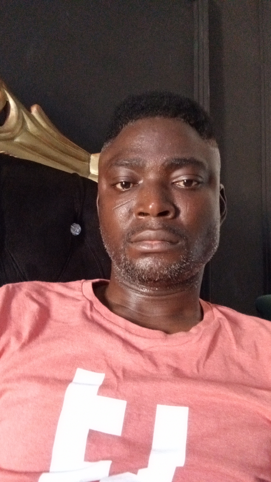 A medium shot depicts a middle-aged Black man wearing a peach-colored t-shirt with a large white graphic. The man has short, dark hair and a salt-and-pepper beard. A faint scar is visible on his right cheek. He is sitting on what appears to be a dark leather chair with gold trim. The background is a dark wall. The location is provided as the city of Sabon Tasha, Nigeria, but there are no details in the image that provide any support for this claim.