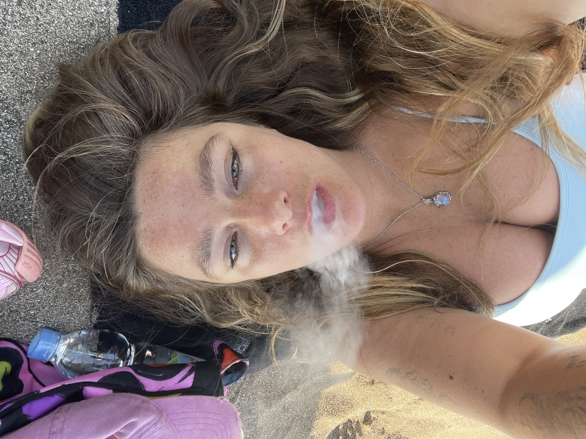 This is a first-person perspective, close-up, overhead shot of a woman lying on a sandy beach. She has long, wavy, light brown hair, with some lighter streaks. Her eyes are open and looking upwards. She is exhaling smoke, which is visible in the air. She is wearing a light blue bikini top and a silver necklace with a teardrop-shaped pendant. There are some freckles visible on her face and shoulders. Her left arm is extended towards the camera, and a tattoo is visible on her forearm.
To the left of the woman, there is a black towel, a pink child's shoe, a plastic water bottle, and a pink towel with a colorful abstract pattern. The ground appears to be a mix of sand and small pebbles. The lighting suggests it is daytime, possibly mid-morning or afternoon, with bright sunlight casting a slight glare on the sand. The overall mood of the image appears relaxed and casual, consistent with a beach setting. There is no visible text in the image.