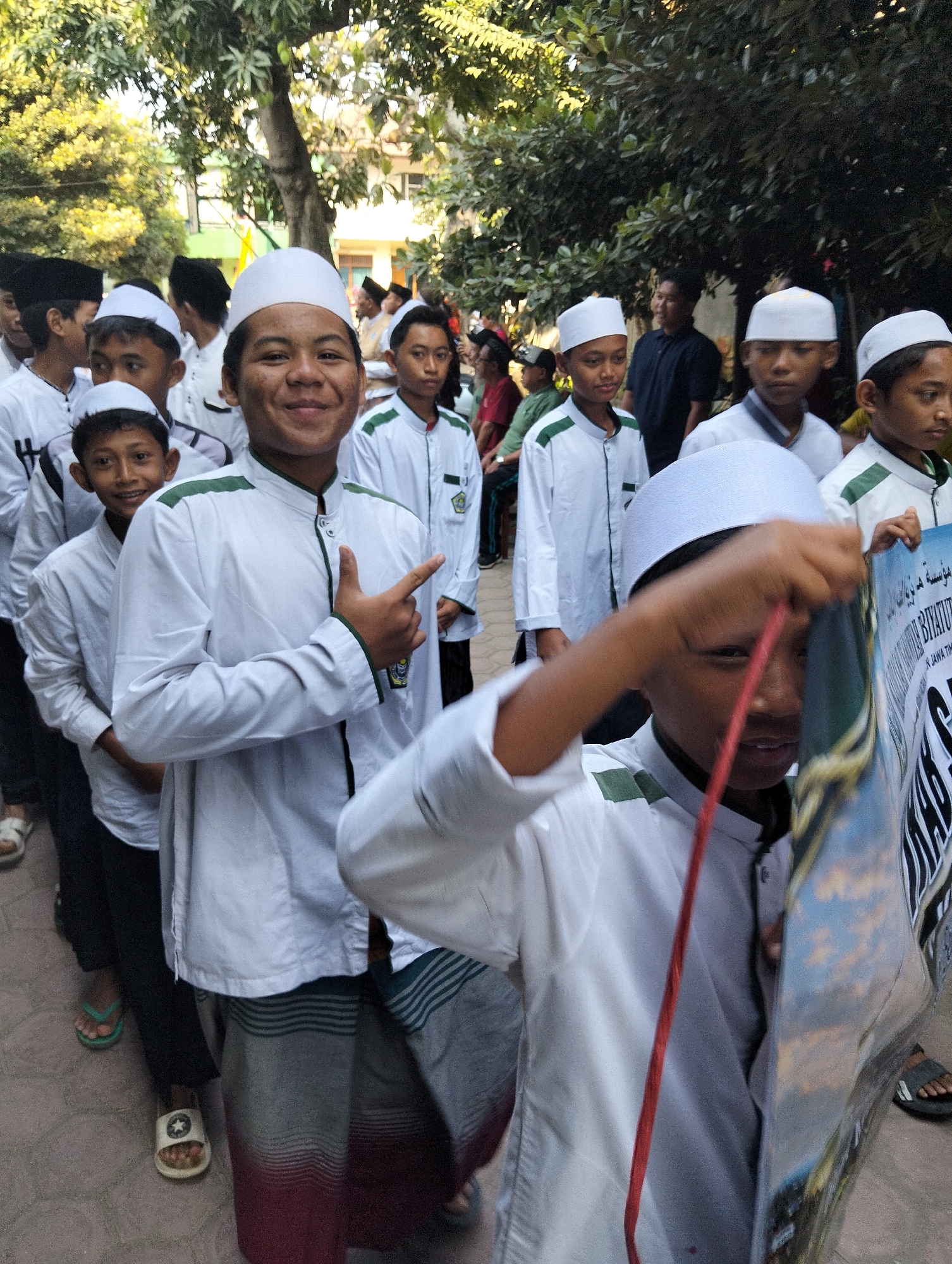 In the city of Kranji, Indonesia, a group of young boys in white robes and skullcaps stand in a line outdoors, some holding a large poster. The boy in the foreground, on the right, is partially obscured by the poster he is holding. He is wearing a white robe and skullcap, and his right hand is visible as he holds the poster. To his left, another boy in a similar outfit smiles at the camera and points with his right hand. Behind them, other boys in white robes and skullcaps are visible, along with some adults in the background. The environment appears to be a street or pathway lined with trees and some buildings in the distance. The lighting suggests it is daytime.