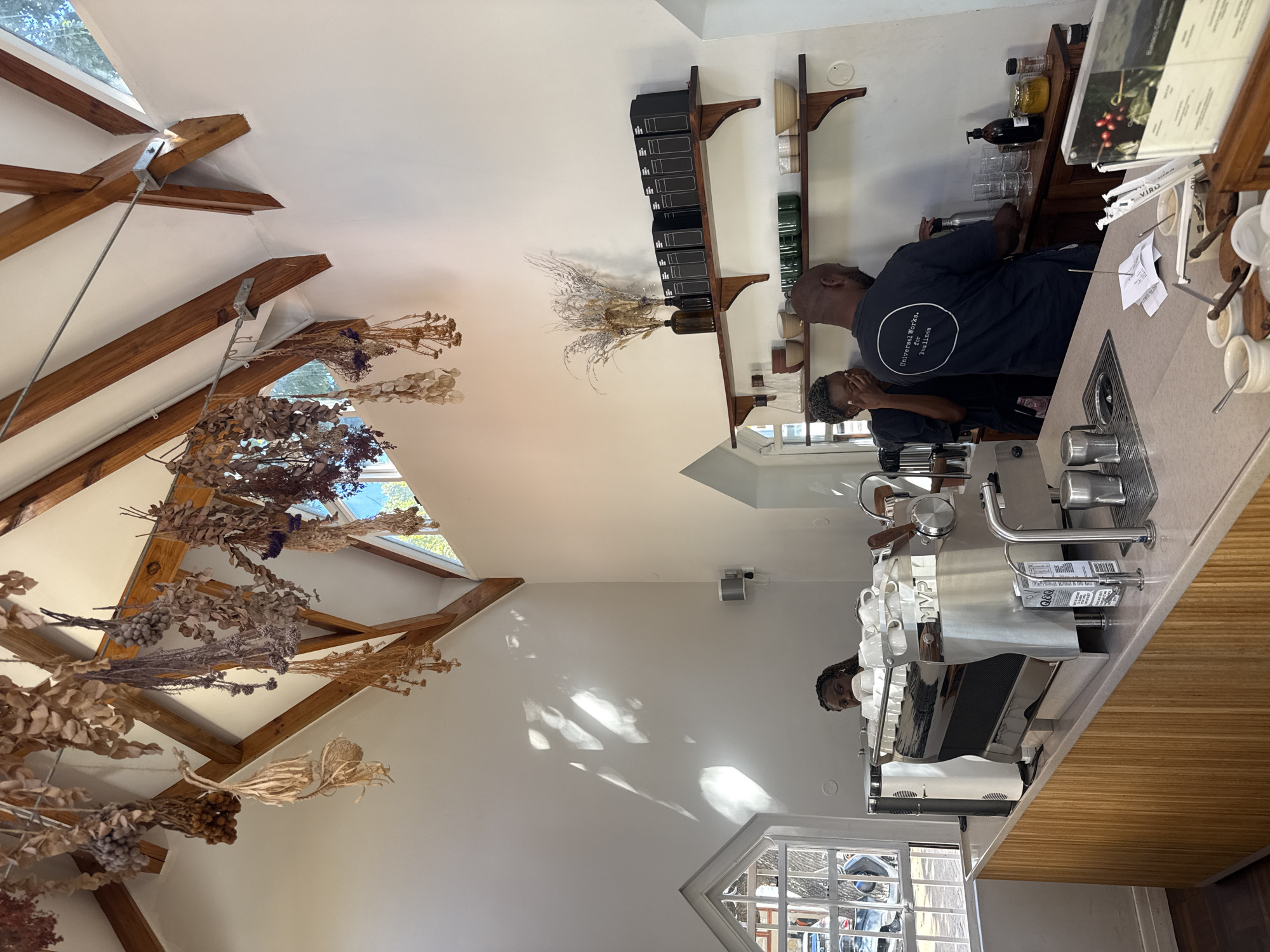 The image depicts the interior of a café, likely in Cape Town, South Africa, given the style of architecture and décor. Two individuals are behind the counter, one is wearing a dark blue t-shirt, and the other has curly hair. They appear to be operating a professional espresso machine, which sits atop a light-colored countertop. Shelves with coffee beans and other items line the wall behind the counter. The café is decorated with dried plants, which hang from a ceiling with exposed wooden beams. Sunlight streams in through windows in the ceiling.