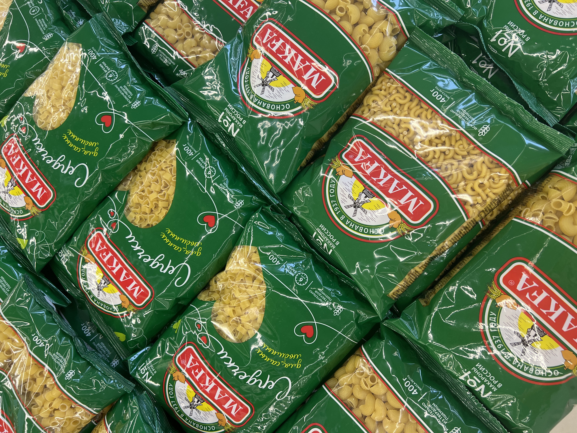 The image displays a close-up, overhead view of multiple packages of Makfa brand pasta, predominantly "Serderki" (small stars) and elbow macaroni. The packaging is a vibrant green with the Makfa logo in red and white, along with Russian text and graphics of wheat stalks and a windmill. Some packages also feature the text "№1 Макароны в России" (No. 1 Pasta in Russia) and "ОСНОВАНА В 1937 ГОДУ" (Founded in 1937). The lighting is bright, suggesting an indoor environment, likely a supermarket aisle or storage area. The arrangement of the packages is layered and slightly staggered, creating a sense of abundance. There are no people or discernable environmental features that would indicate a specific location within Chaikovsky, Russia, beyond the brand being Russian in origin.