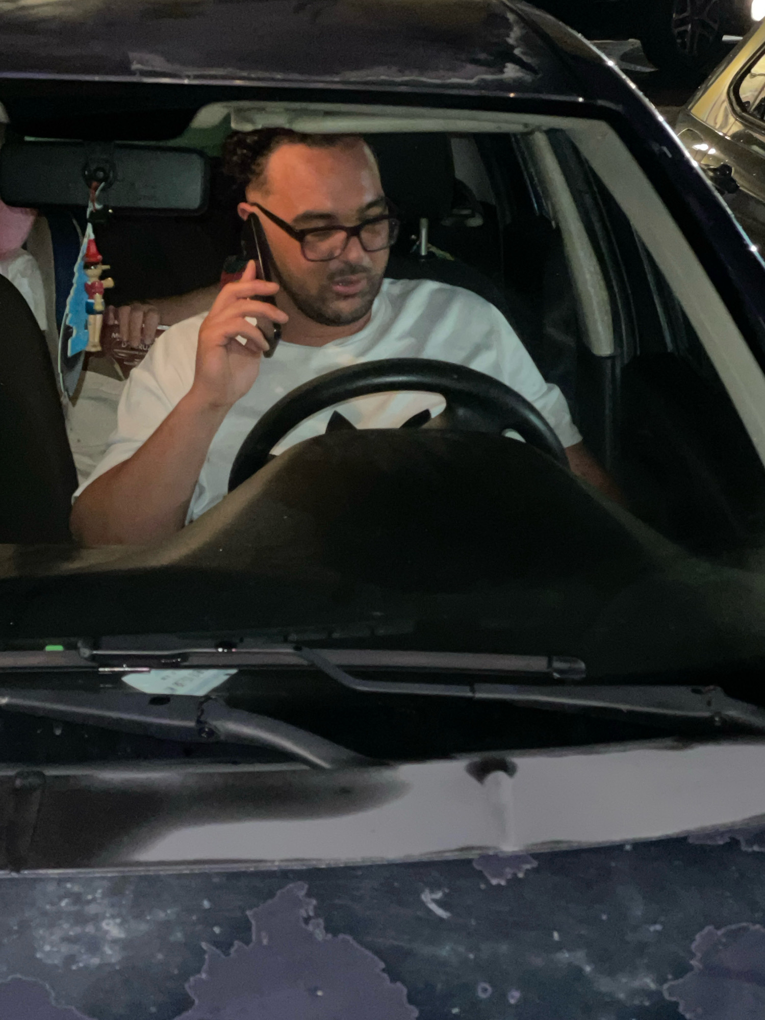 A man with dark, curly hair and glasses is in the driver's seat of a car, talking on a black cell phone. He is wearing a white t-shirt. A red and blue air freshener shaped like Pinocchio hangs from the rearview mirror. Another person is partially visible in the passenger seat. The scene takes place in Altopascio, Italy.