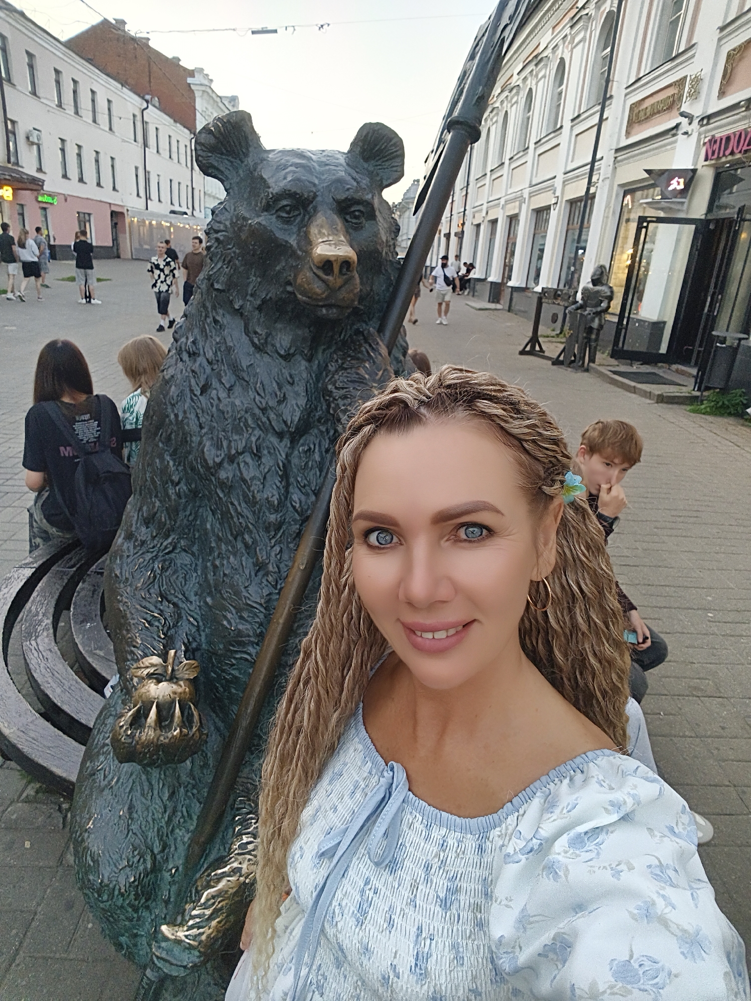 A woman with blonde braided hair and blue eyes takes a selfie in front of a bronze bear statue on a city street. She is wearing a light blue off-the-shoulder top with a floral pattern. Behind her, the bear statue holds a large spear-like object. Several other people are visible in the background, including children and adults walking along the street and sitting on a bench. The street is paved with stone and lined with buildings with traditional architecture. This scene is located in Yaroslavl, Russia, likely on the pedestrian street of Kirov.