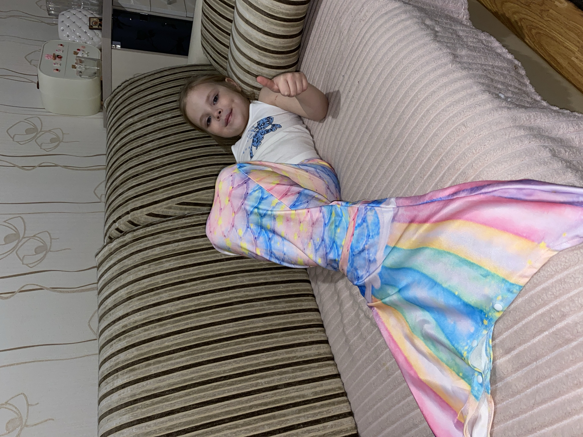 A young girl with light-colored hair is captured lying on her side on a couch, smiling cheerfully and giving a thumbs-up gesture towards the camera. She exudes a happy and playful emotion.
She is dressed in a white tank top featuring a stylized blue graphic design on the front. Her lower body is covered by a vibrant, rainbow-colored mermaid tail, which appears to be a costume or a blanket. This mermaid tail showcases pastel shades of pink, yellow, blue, and purple, with a subtle scale-like pattern, and has white snap buttons visible along one side near the fin.
The setting is indoors, likely a living room or a child's space. The couch she rests on has two sections: one upholstered with horizontal brown and light beige/cream stripes, and the other covered in a light, ribbed, possibly corduroy-textured fabric. In the background, to the left of the girl, a light-colored wall features subtle, wavy, abstract patterns. Further back, a white decorative box or organizer topped with various small, colorful items is visible on a surface, next to what appears to be a cabinet or shelf with reflective elements. The lighting suggests an indoor environment, possibly illuminated by artificial light. There is no discernible text visible in the image. The location context provided is Votkinsk, Russia.