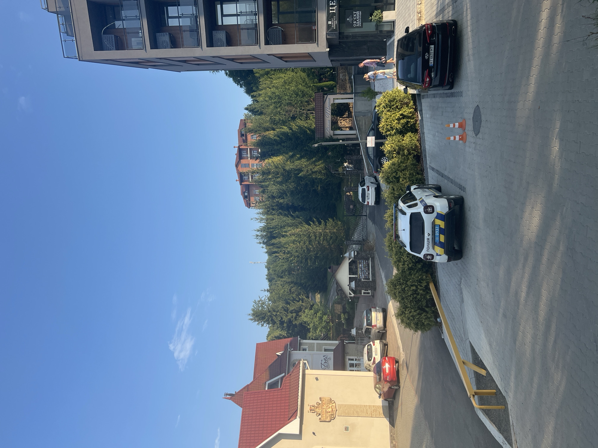 The image shows a parking lot in front of a modern building in the city of Сонячне, Ukraine. A white police car with the word "ПОЛІЦІЯ" on its side is parked in the foreground. To its right is a black sedan and two orange traffic cones. In the background, there is a multi-story building with balconies and a smaller building with a red roof. A wooded hill with a large residential building on top is visible behind the buildings. The sky is clear and blue, indicating good weather. Two people are walking on the sidewalk next to the modern building.