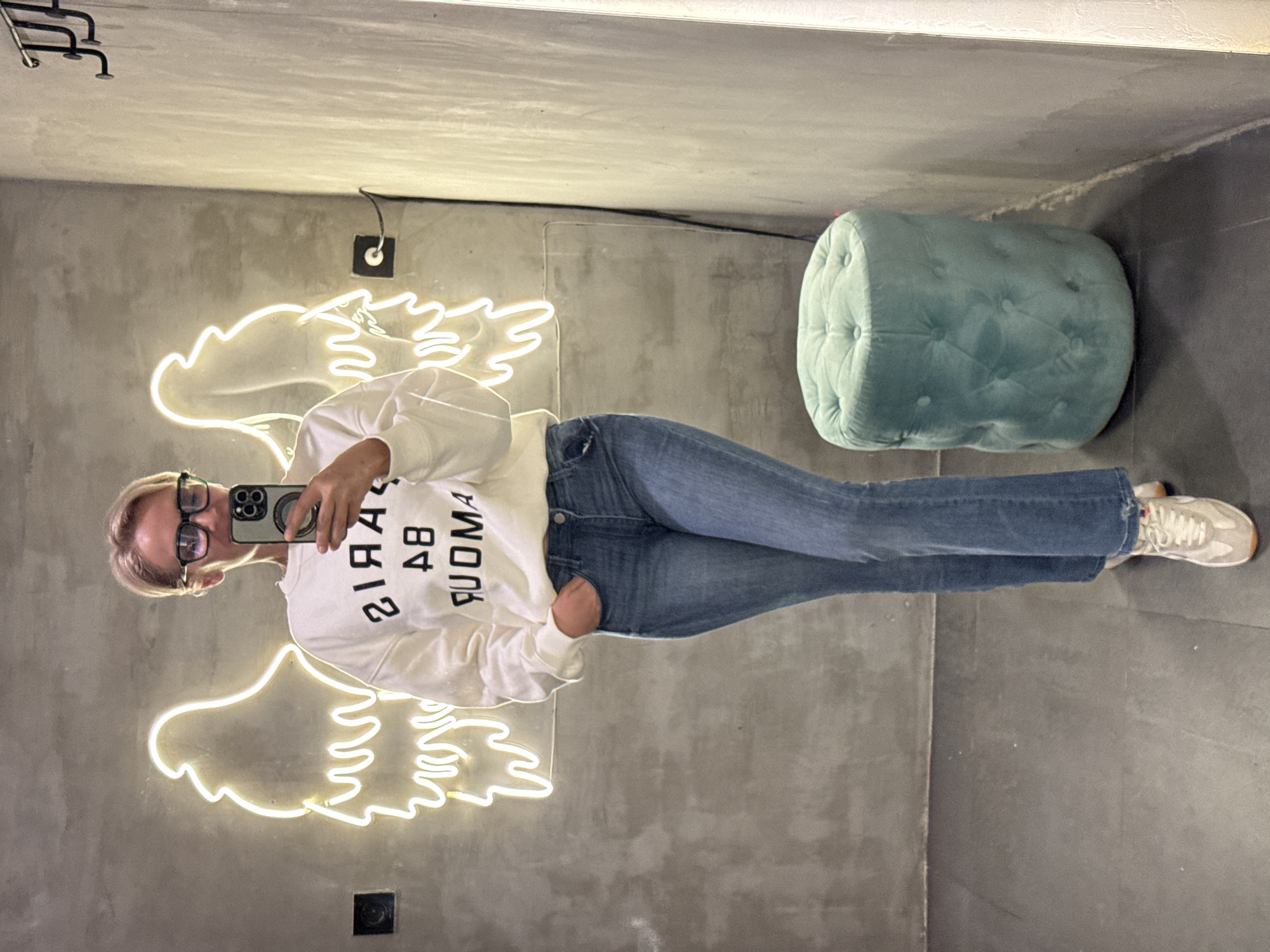 A woman stands in a room with a concrete wall, illuminated by a neon angel wing sign. She wears glasses, a white sweatshirt with the text "Paris AB Ruoma" printed on it, jeans, and sneakers. She holds a phone in her hand, presumably taking a selfie in front of a mirror. To the right of her, there is a light turquoise ottoman. The location is Kuta Selatan, Indonesia.