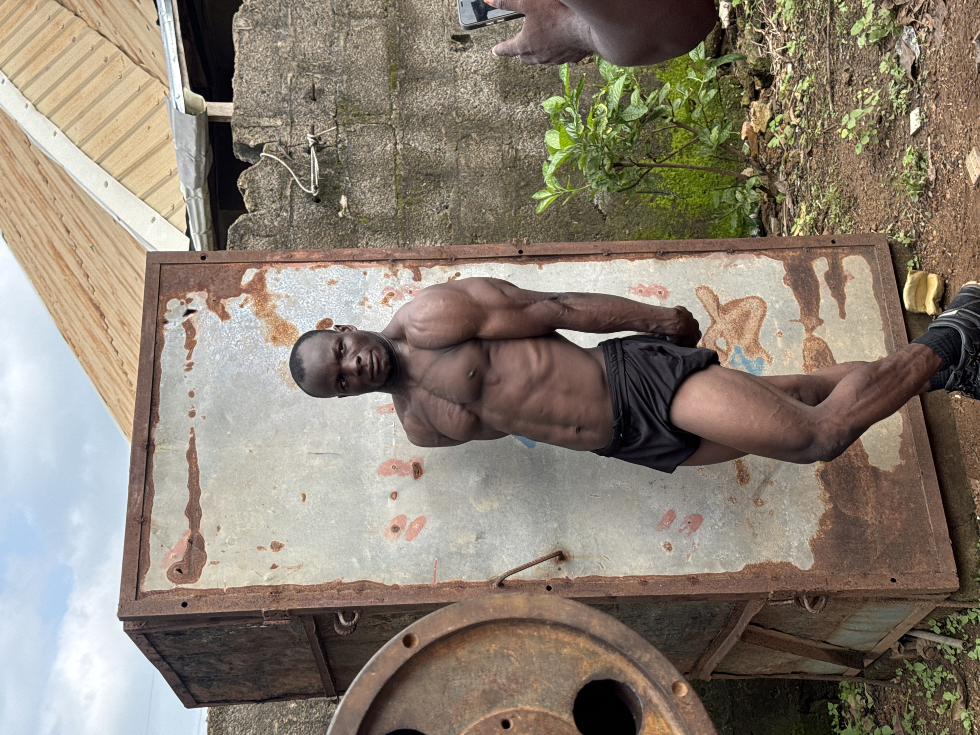 An African man with well-defined muscles stands in front of a rusty metal structure. He is wearing black shorts and dark shoes. The man's posture suggests he is posing for a photo, as another person's hands are visible on the right side of the frame, holding a smartphone, likely taking a picture.
The background includes a rough-textured concrete wall with some green moss, a corrugated metal roof of a building, and a small green plant. The ground is covered with dirt and some scattered debris. The overall environment appears to be outdoors, possibly in a yard or an industrial-looking area. The presence of the rusty metal structure and the rough wall suggests an urban or semi-urban setting, consistent with locations in Akwanga, Nigeria. The lighting indicates it is daytime, with a cloudy sky visible.