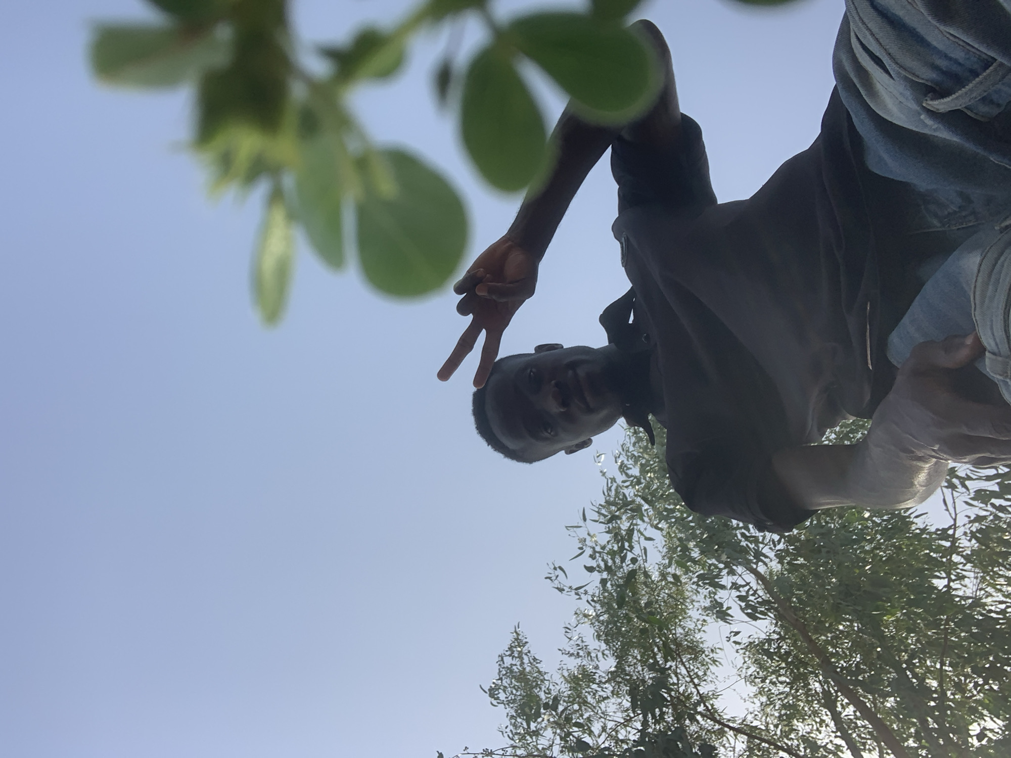 A low-angle shot depicts a man with dark skin, viewed from below, making a peace sign with his right hand. He is wearing a dark shirt and blue jeans. Above him, on the upper left, are green leaves of a tree, slightly out of focus. In the background, below the man, are trees with green foliage against a clear, light blue sky. The image appears to have been taken outdoors, potentially in Gbeleko, Nigeria, as specified.