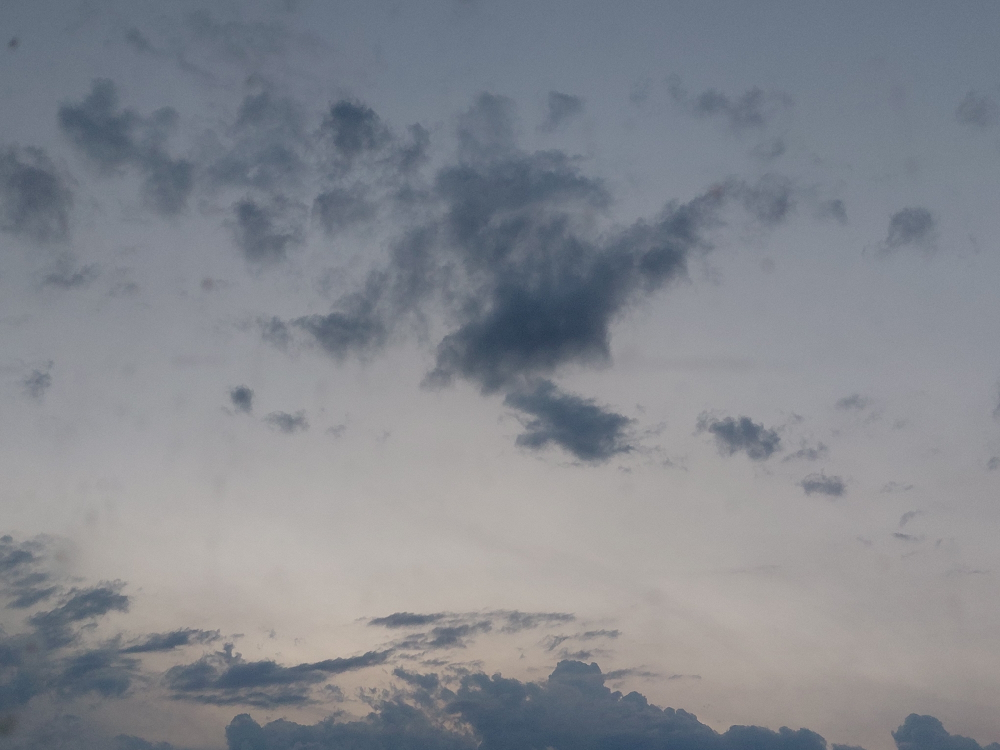 The image is a photograph of a sky filled with clouds. The clouds are varied in size and density, ranging from small, wispy formations to larger, more defined masses. The sky is overcast, with a muted, grayish-blue hue. There is a subtle gradient in the sky, with the lower portion appearing lighter and brighter, suggesting the presence of sunlight filtering through the clouds. The overall mood of the image is somber and atmospheric. There are no discernable objects or people in the image, and it is an abstract depiction of the sky. The location is listed as Unknown, Unknown, indicating that the specific geographical context of the photograph is not provided.