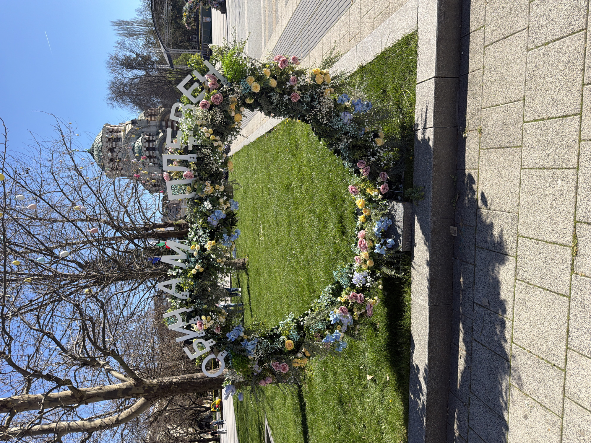 The image captures a floral archway set up on a patch of grass next to a paved walkway. The arch is adorned with a variety of flowers, including pink roses, yellow blooms, and blue hydrangeas, interspersed with greenery and baby's breath. White Cyrillic letters are arranged along the arch, spelling out a phrase.
In the background, the dome of a building is visible, and trees with bare branches stand to the left. Decorations are hung on the branches of the trees, resembling Easter eggs.
The scene is located in Pleven, Bulgaria, and the building in the background appears to be the St. George the Victorious Chapel Mausoleum, a notable landmark of the city. The weather seems sunny and clear, with a blue sky visible above. No people are clearly visible within the arch.