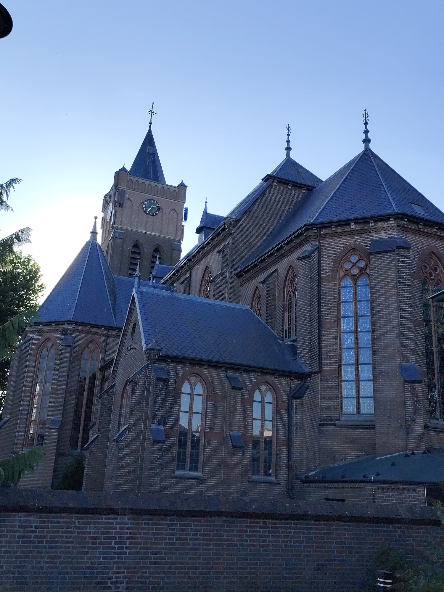 This is a medium-angle, exterior shot of the Roman Catholic Church of Saint Anthony of Padua (Sint-Antonius van Paduakerk) in Schaijk, Netherlands. The church is constructed of dark brick and features a tall, square tower with a clock face. The tower is topped with a pointed, dark blue roof with a spire and cross. The church also has several other pointed, dark blue roofs with spires. Ornate, arched windows with stained glass are visible on the main body of the church. The church is partially obscured by trees on the left side of the image. A brick wall runs along the bottom of the frame, in front of the church. The sky is clear and blue. The sun is shining from the upper left, casting a slight glare on the clock face and silhouetting some of the church's architectural features. There are no people visible in the image.