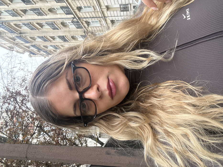 A selfie-style portrait depicts a woman in an urban outdoor setting identified as Paris, France, due to distinctive architectural elements. The woman, centrally framed, has long, wavy blonde hair with a middle part. She wears dark-framed rectangular eyeglasses and a dark grey, ribbed, long-sleeved top featuring a central half-zip and a white embroidered 'SKIMS' logo with a checkmark symbol on the upper left chest. Her expression is neutral, and her gaze is directed towards the camera. A manicured hand is partially visible at the bottom right, appearing to touch her hair.
The background consists of a multi-story Haussmannian building on the right, characterized by its light-colored stone facade, repetitive window patterns, and black wrought-iron balconies. On the left, the branches of a deciduous tree with sparse, reddish-brown leaves are visible, alongside a dark, vertical pole. The sky is overcast and light grey. A light-colored vehicle is dimly visible in the distant street scene.